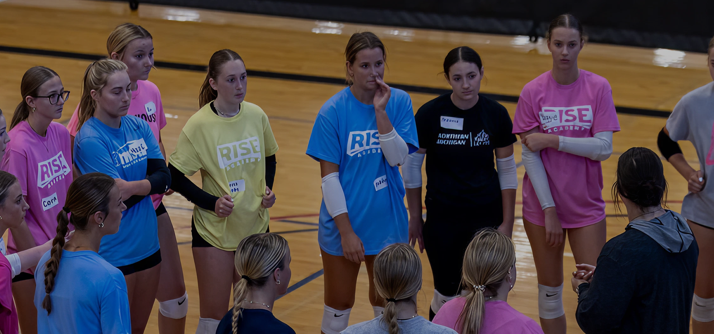 A group of female volleyball players wearing colorful jerseys with 'RISE' printed on them, standing in a gymnasium, listening intently to a coach.
