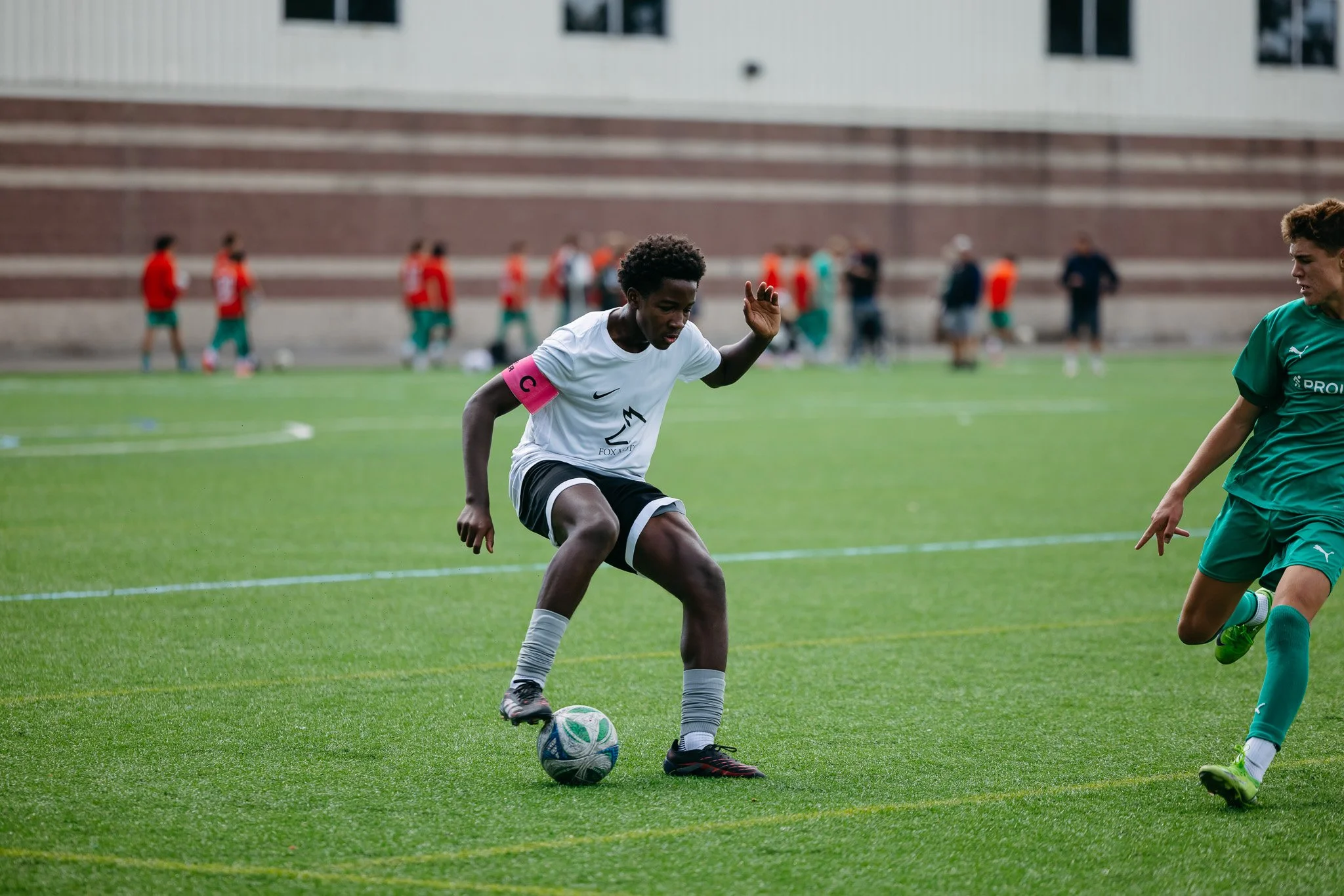 A soccer player in a white uniform with a pink captain's armband is kicking a soccer ball on the field during a game, with other players and spectators in the background.