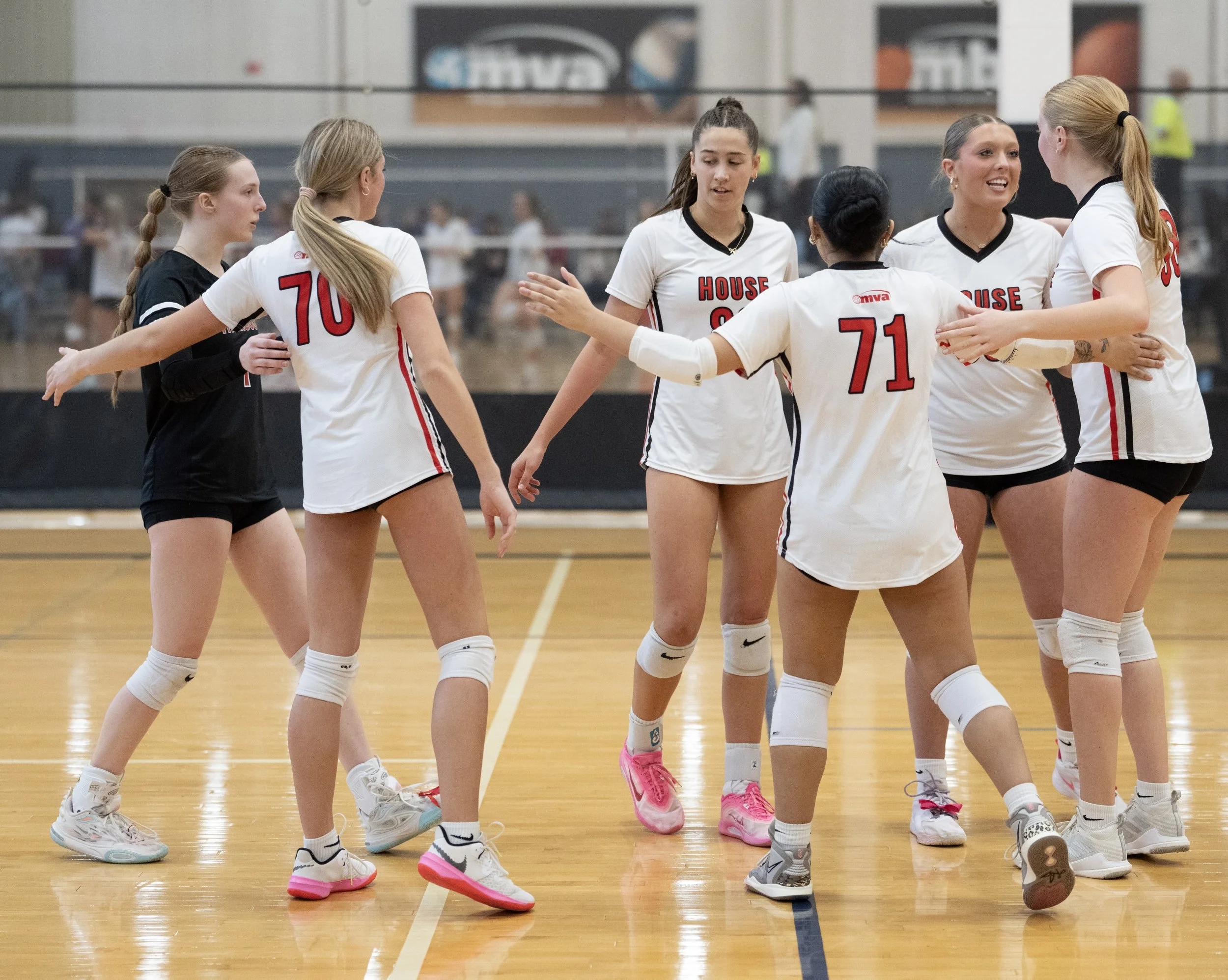 Seven female volleyball players in uniforms are gathered in a circle on an indoor court, engaged in conversation during a break or timeout.