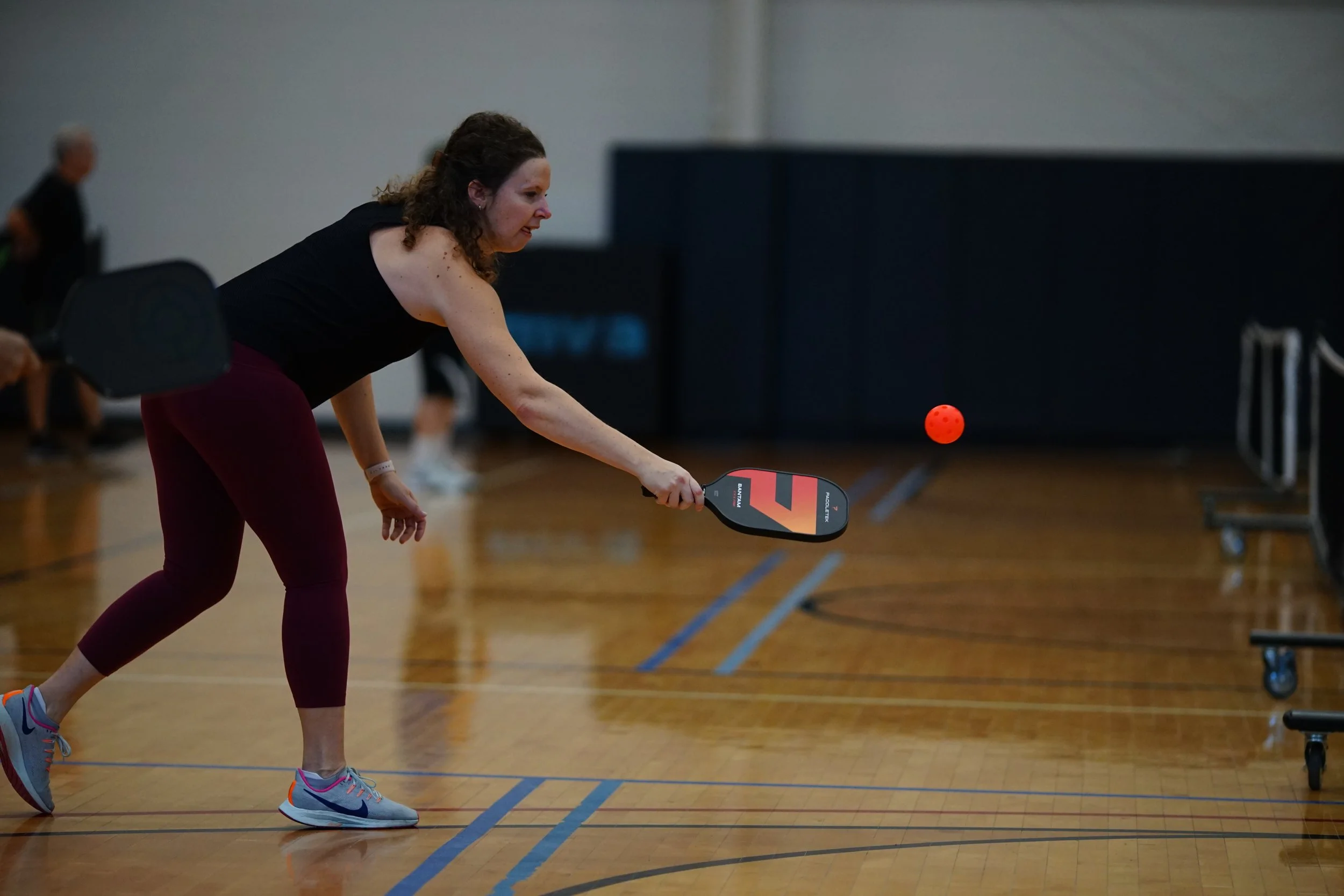 A woman playing pickleball in an indoor gym, preparing to hit an orange ball with a paddle.