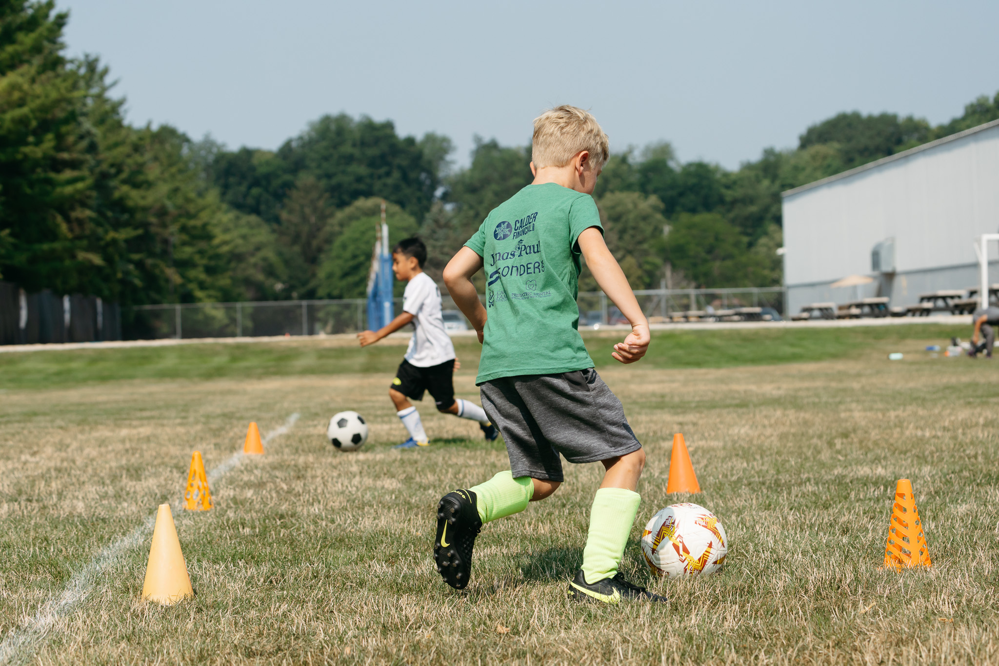 Young boy practicing soccer dribbling drills on a field, with cones set up for training, and other children visible in the background.