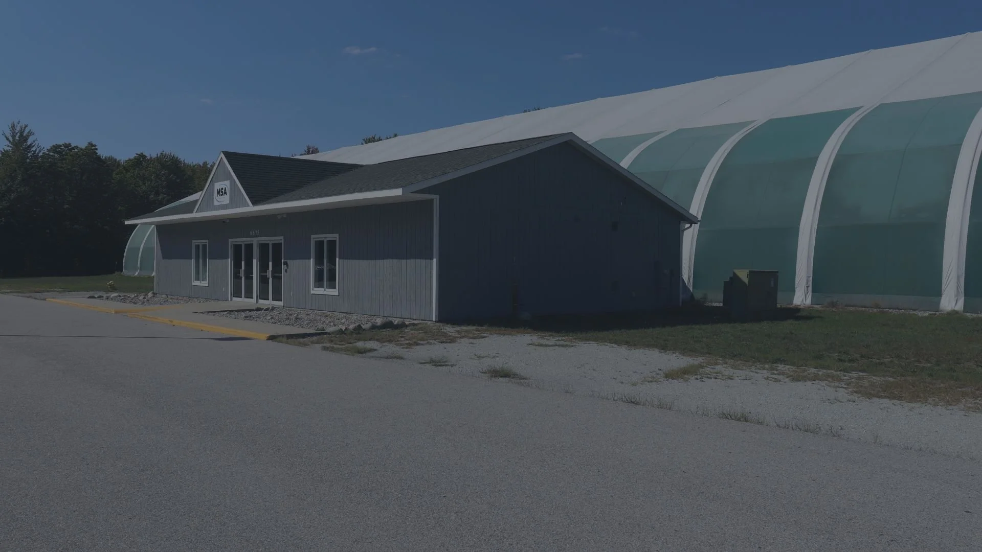 Small gray building with a dark roof and glass door, located next to a large white greenhouse with a translucent curved roof.