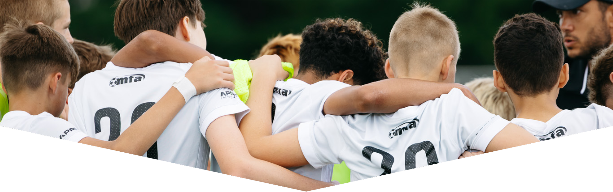 A group of young boys in white sports jerseys huddled together in a circle, likely during a team discussion or prayer before a game, outdoors on a sports field.