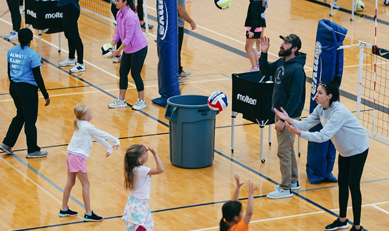 Children playing volleyball in an indoor gym, supervised by coaches, with spectators watching.