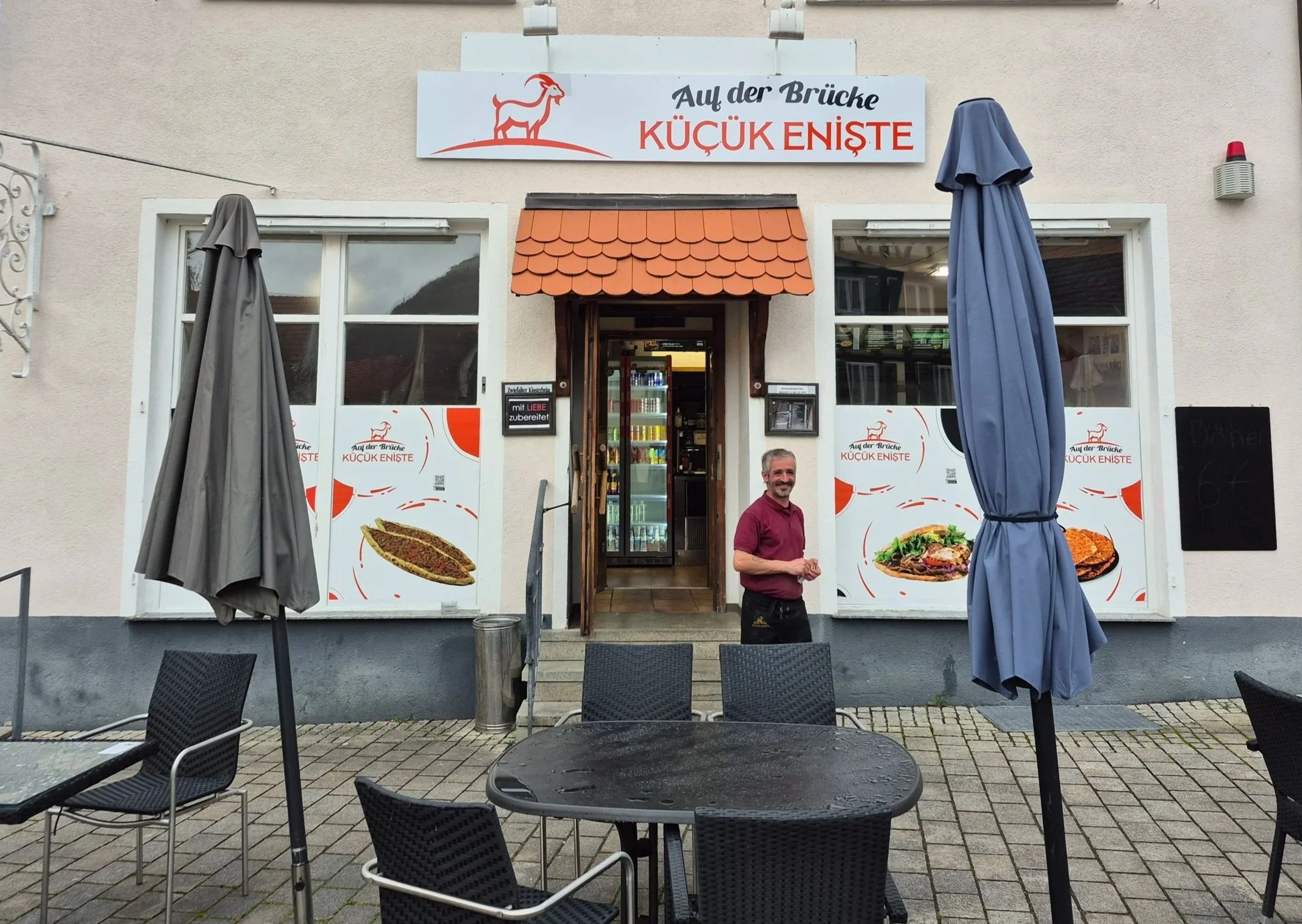 Außenansicht eines Restaurants mit Tischen und Stühlen, einem Mann vor der Tür, drei eingepackte Sonnenschirme, und Speisebilder an den Fenstern.