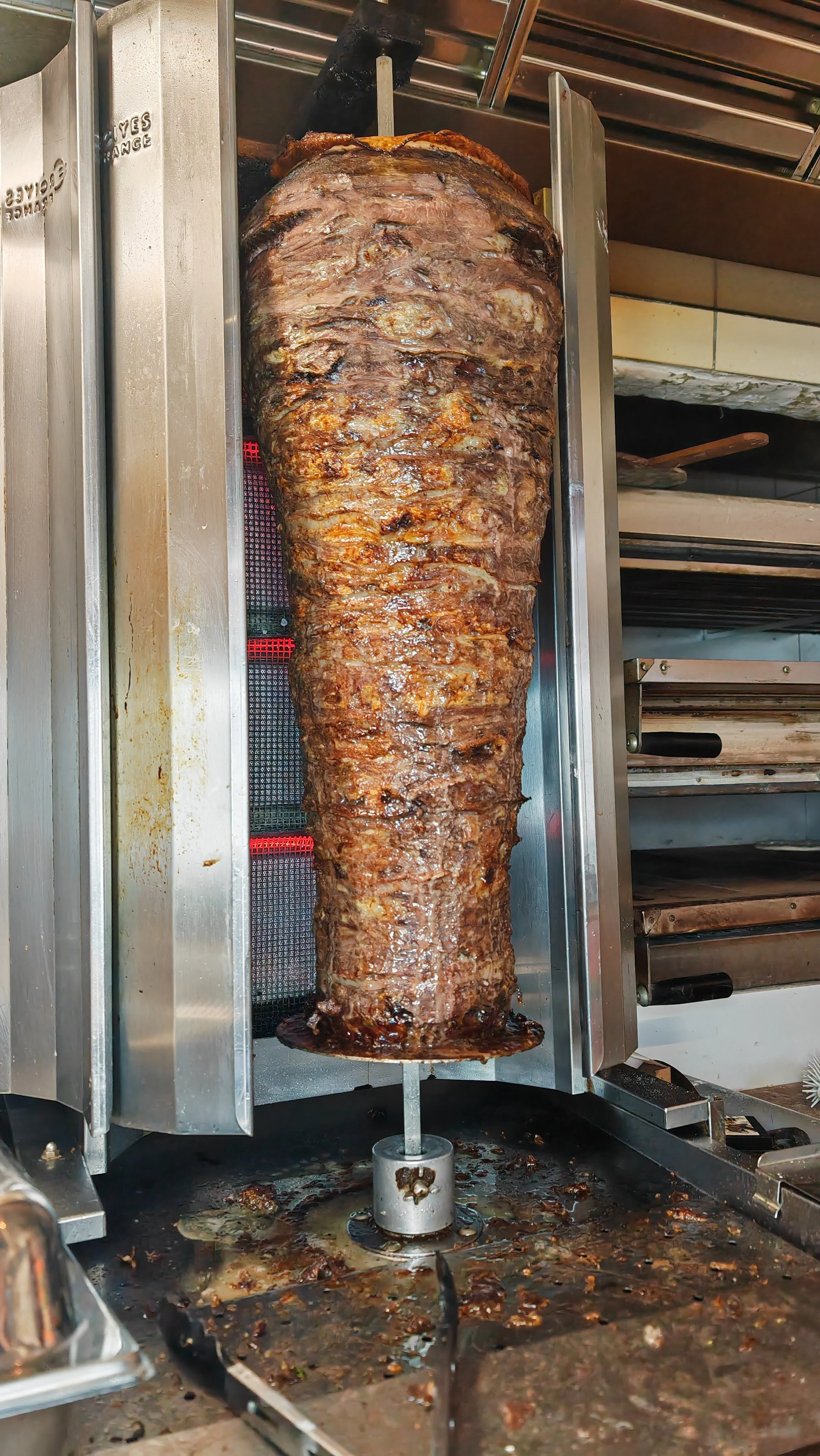 Großer Döner-Spieß mit gebratenem Fleisch auf einem Drehstativ in einer Küchenumgebung.
