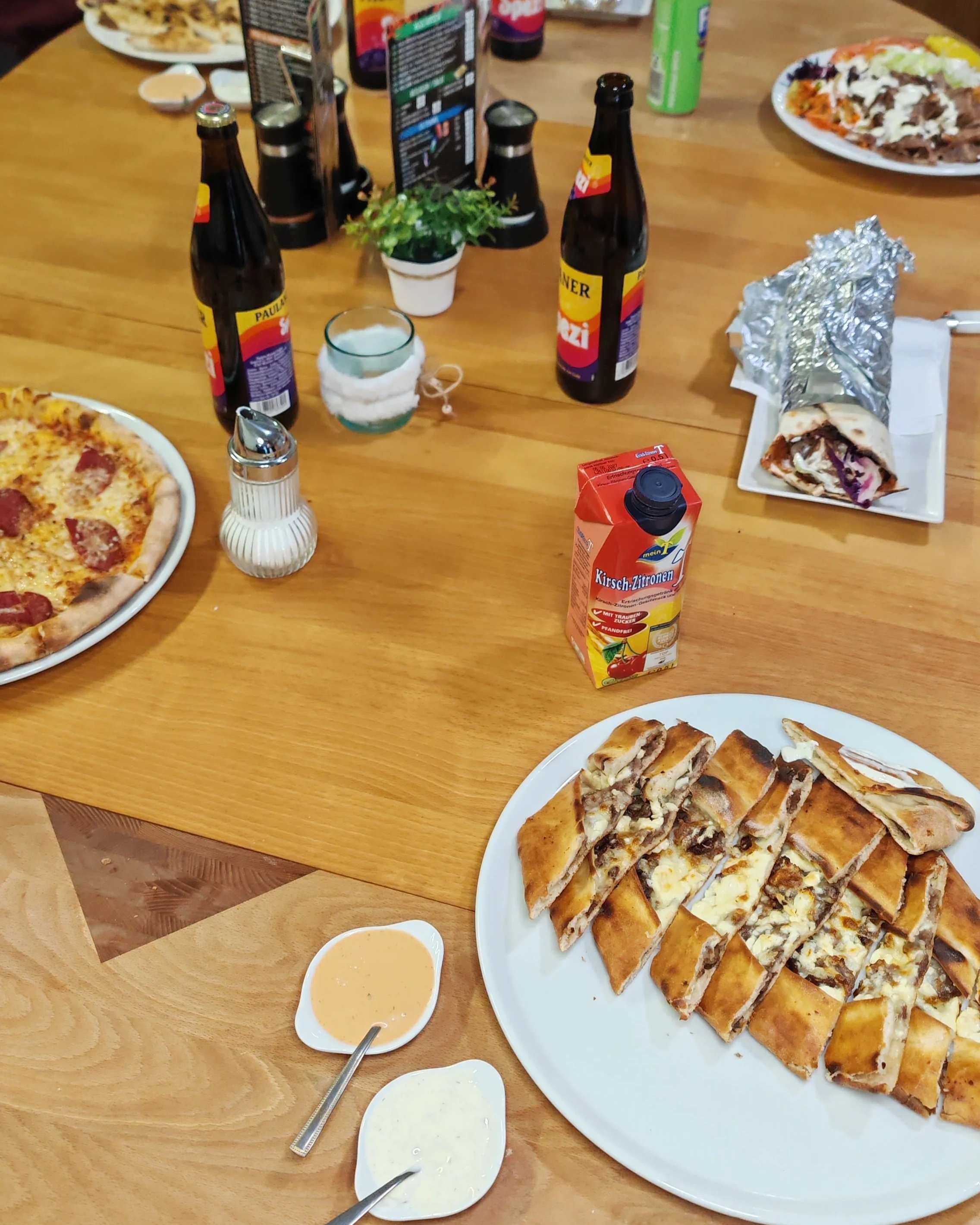 Ein Esstisch mit verschiedenen Speisen und Getränken, darunter eine Pizza, gefüllte Teigtaschen, verschiedene Flaschen Bier, eine Packung Kirsch-Zitronen Saft, ein Burrito, kleine Schälchen mit Soßen, eine Salatplatte und eine kleine Topf mit Pflanzen.