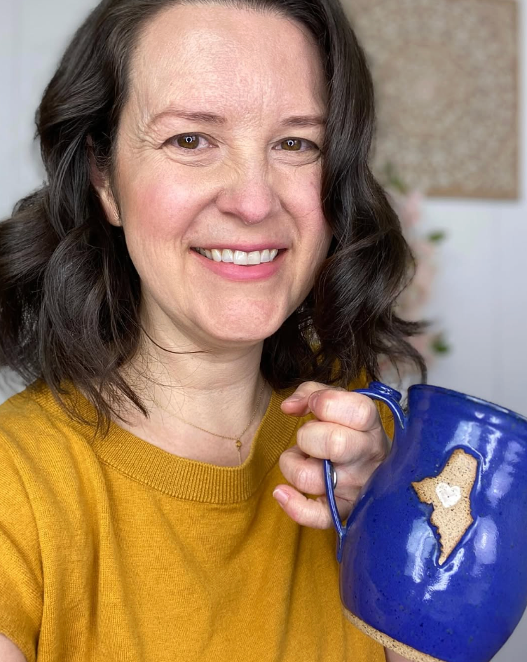A woman with shoulder-length brown hair smiling and holding a blue mug with a cookie shaped like the state of Minnesota with a heart in the center, wearing a yellow shirt.
