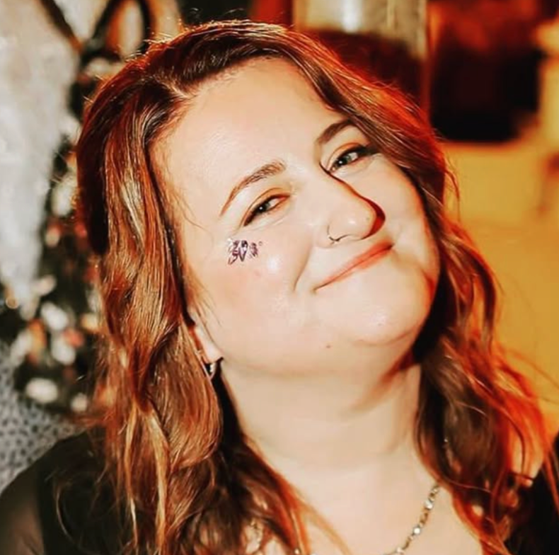 A woman with red hair smiling and making a playful face, wearing a nose ring and an earring, with a tulip tattoo on her cheek.