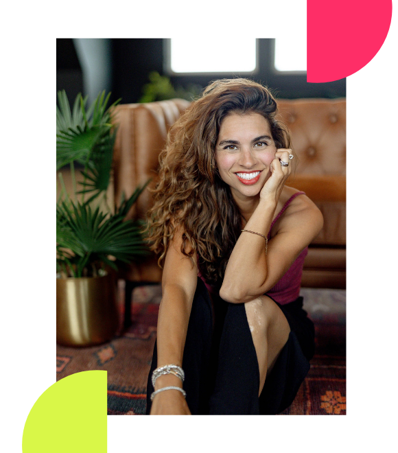 Lisa Vee, host of  The Six-Figure Doula Podcast,  sitting on the floor in a stylish living room, smiling at the camera, wearing a sleeveless top and black pants, with green plants and a brown leather couch in the background.