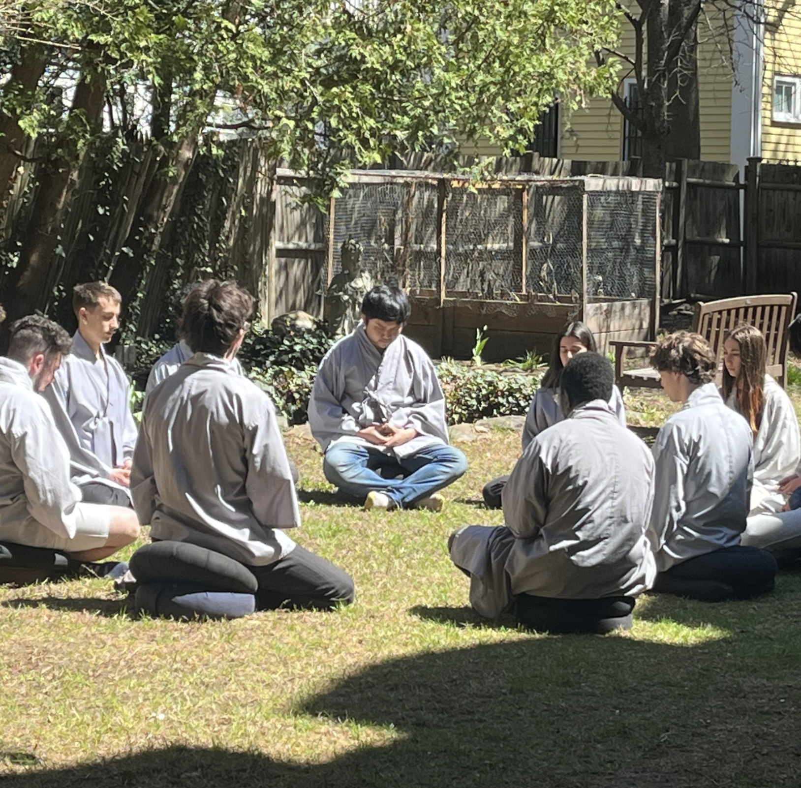 A group of people sitting cross-legged on the grass in a circle, some on meditation cushions, in a backyard with trees, a wooden fence, and a small garden area.