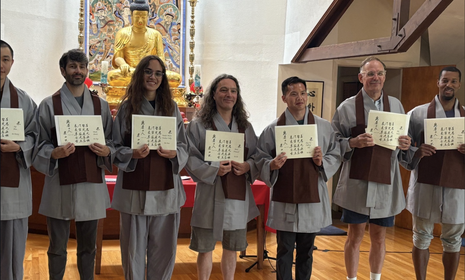 Group of seven people dressed in traditional attire standing in front of a Buddha statue holding certificates.