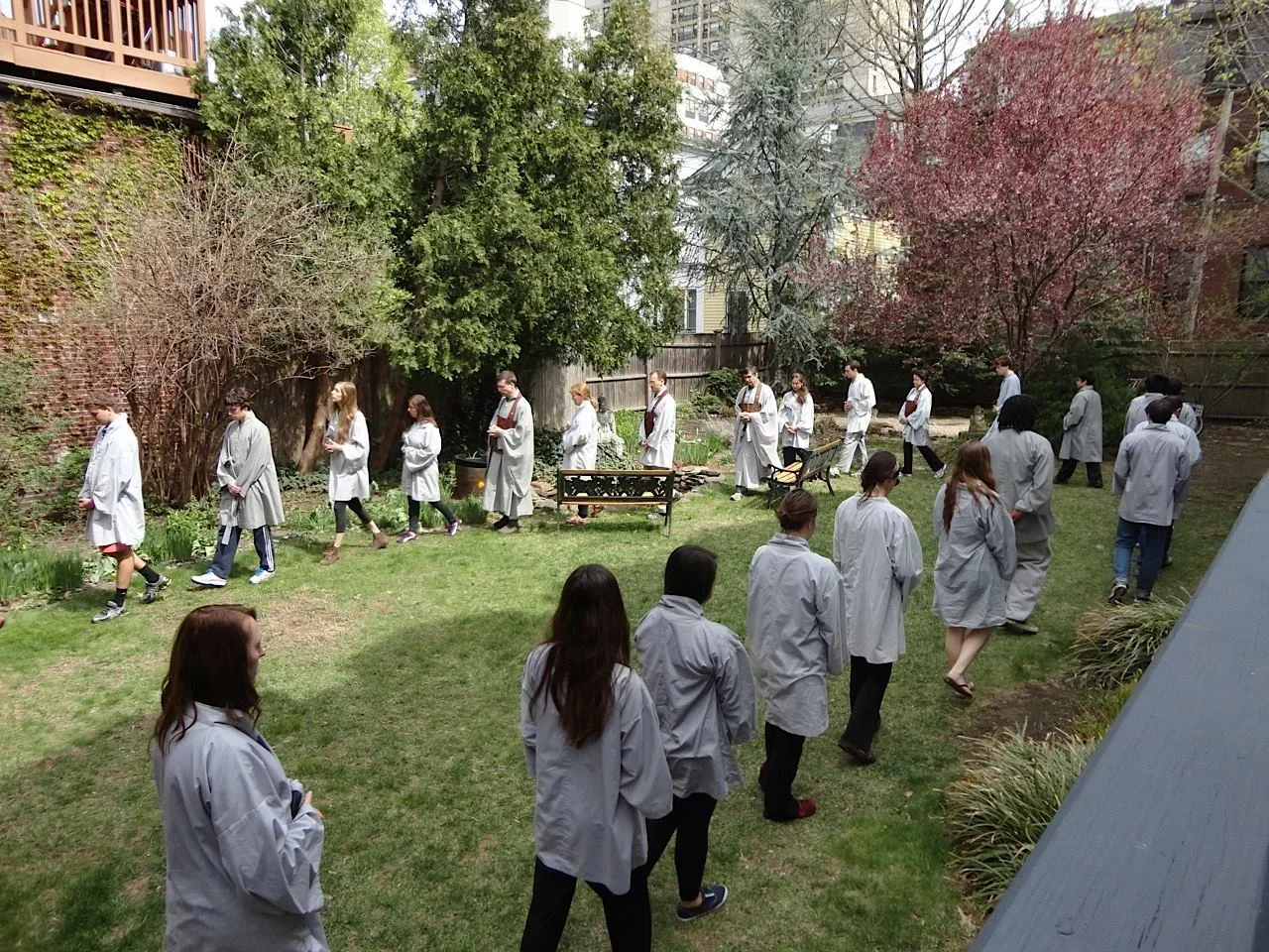 Group of people dressed in white coats walking in a line in a backyard garden with trees and bushes.