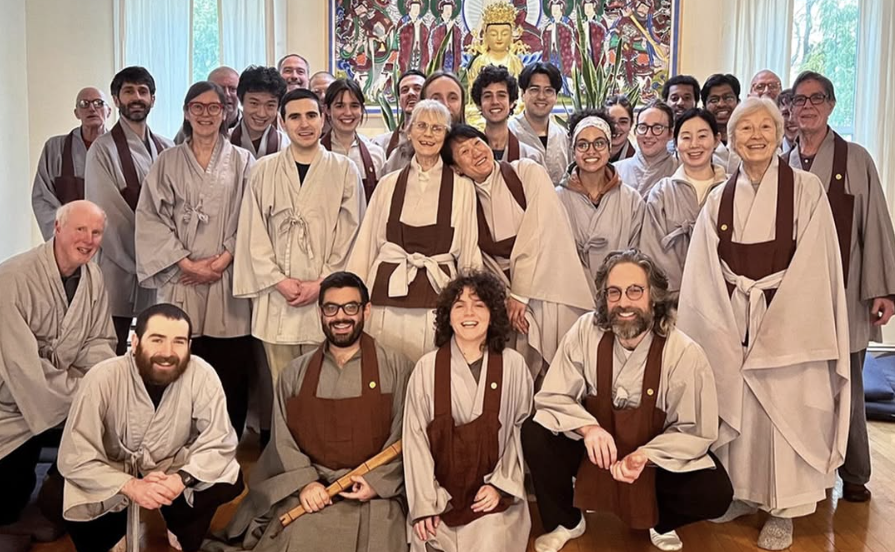 A group of people dressed in traditional robes, gathered together indoors for a group photo in front of a Buddha statue and colorful artwork.