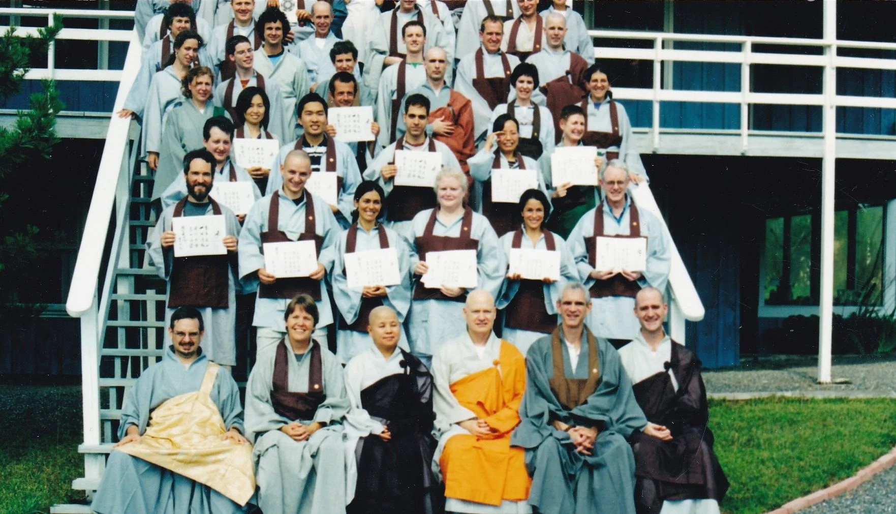 Group of people dressed in traditional robes, some holding certificates, posing on stairs outside building, smiling for a graduation photo.
