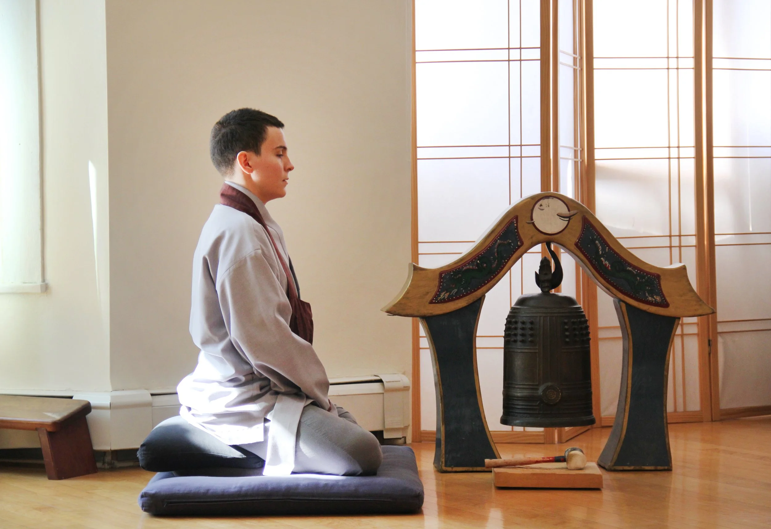 A young person with short dark hair in traditional clothing kneeling on a cushion, facing a bell with a wooden frame decorated with a moon face. The setting appears to be a peaceful, well-lit room with wooden and paper screens.