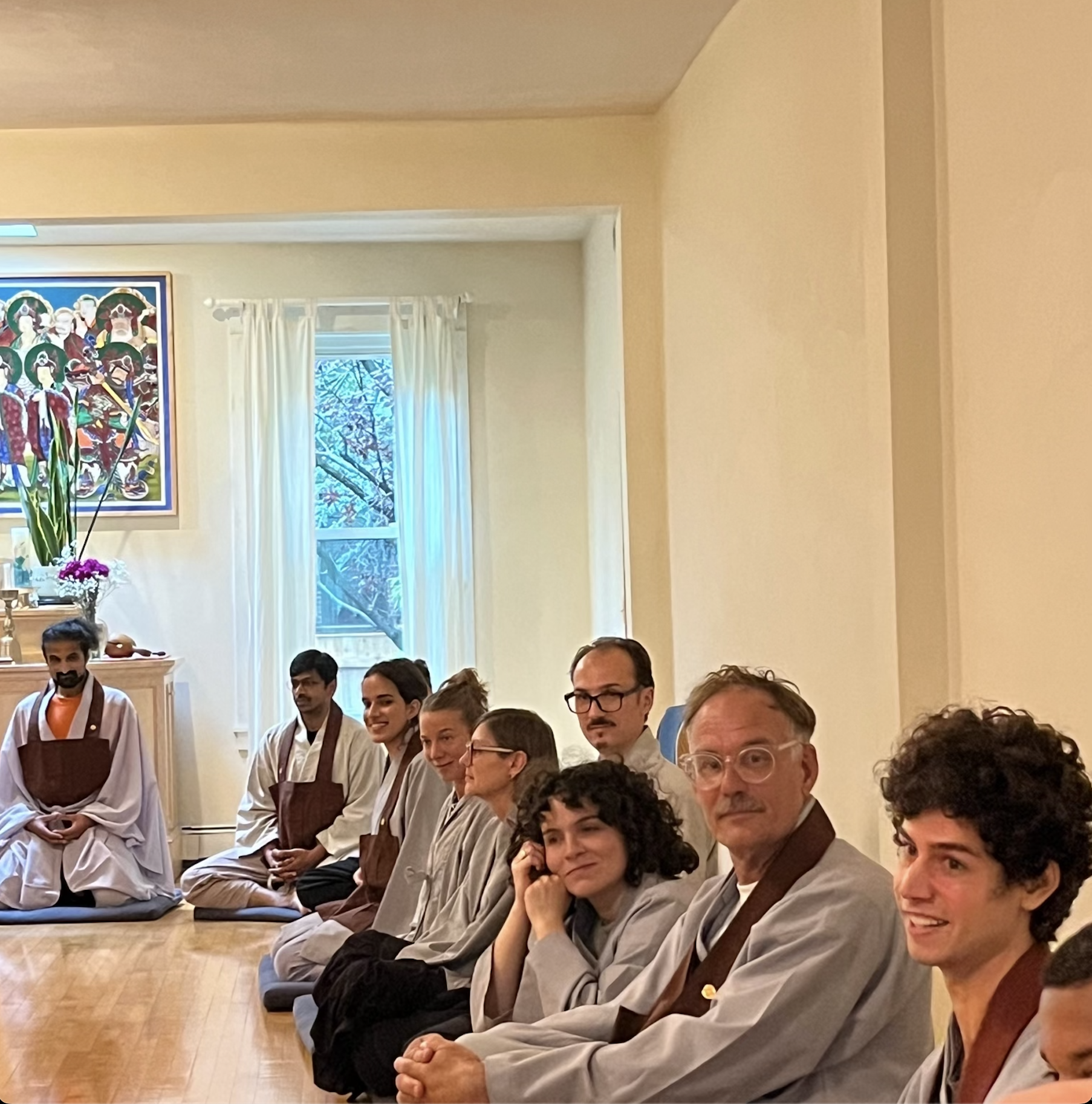 Group of people sitting in a room, some wearing traditional robes, with a burning incense and a colorful painting on the wall.