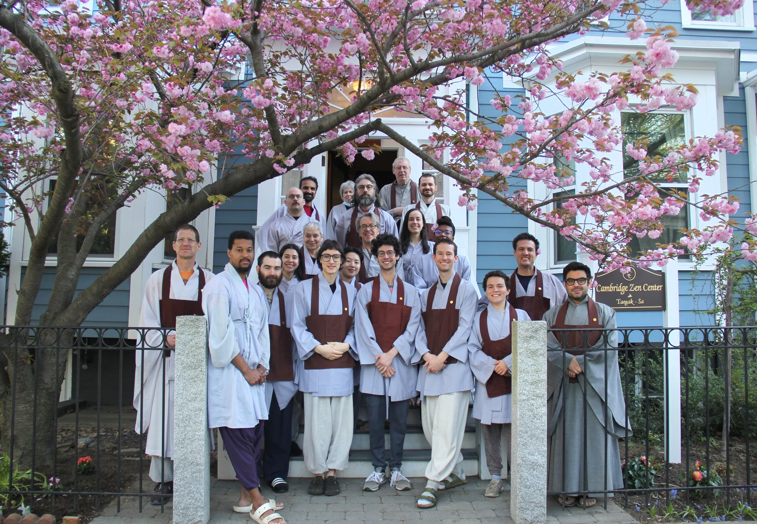Group standing in front of CZC front door wearing grey robes and brown kasas under a blooming cherry tree