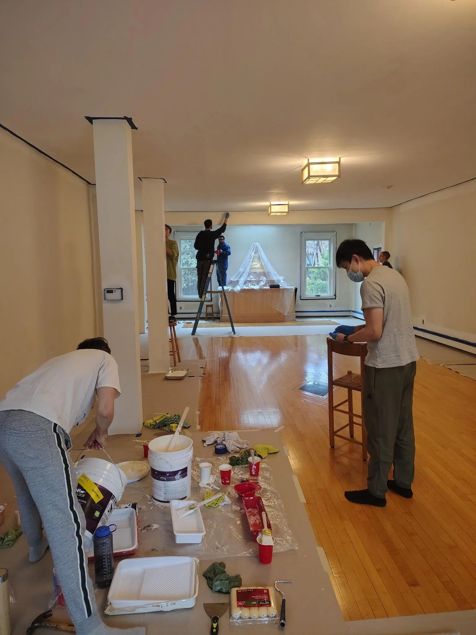 People working and painting inside a room with hardwood floors, some wearing masks, and a table covered with painting supplies and tools, with a window and a covered object in the background.