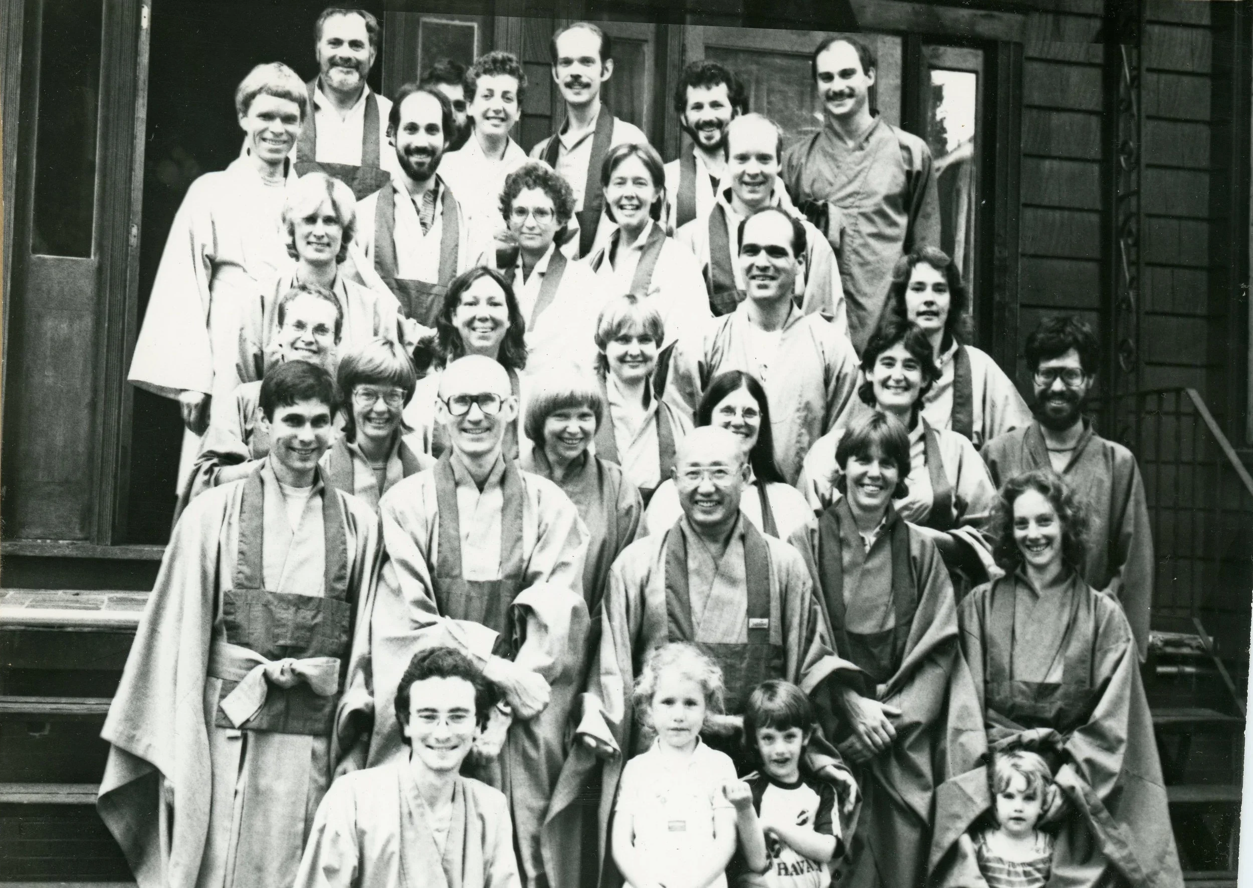 Black and white group photo of adults and children standing on the steps outside a building, some in traditional robes, smiling at the camera.