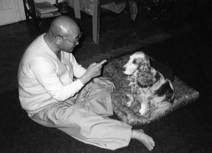 A man sitting on the floor using a smartphone next to a sitting dog on a blanket.