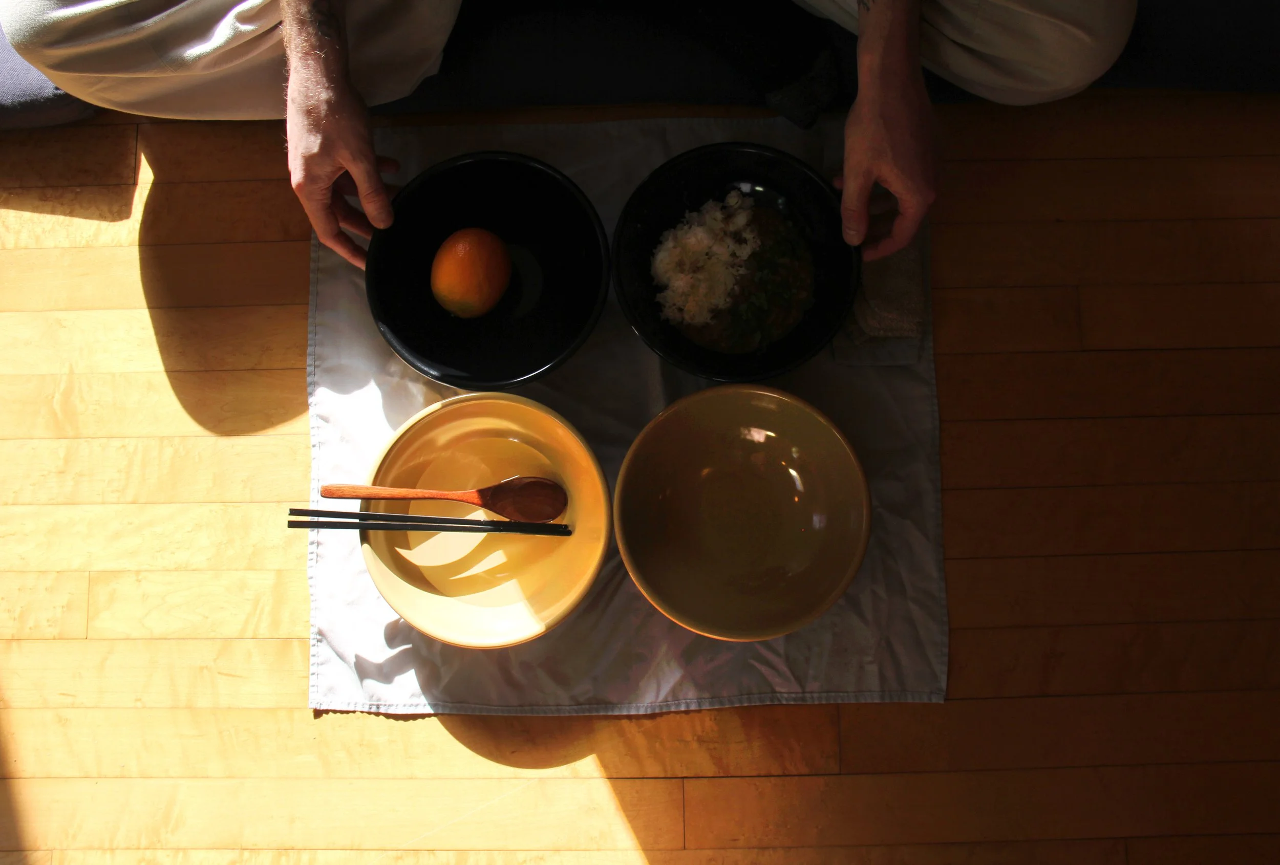 Hands placing black bowls on a cloth-covered table with a yellow bowl, empty brown bowl, and utensils, in sunlight.