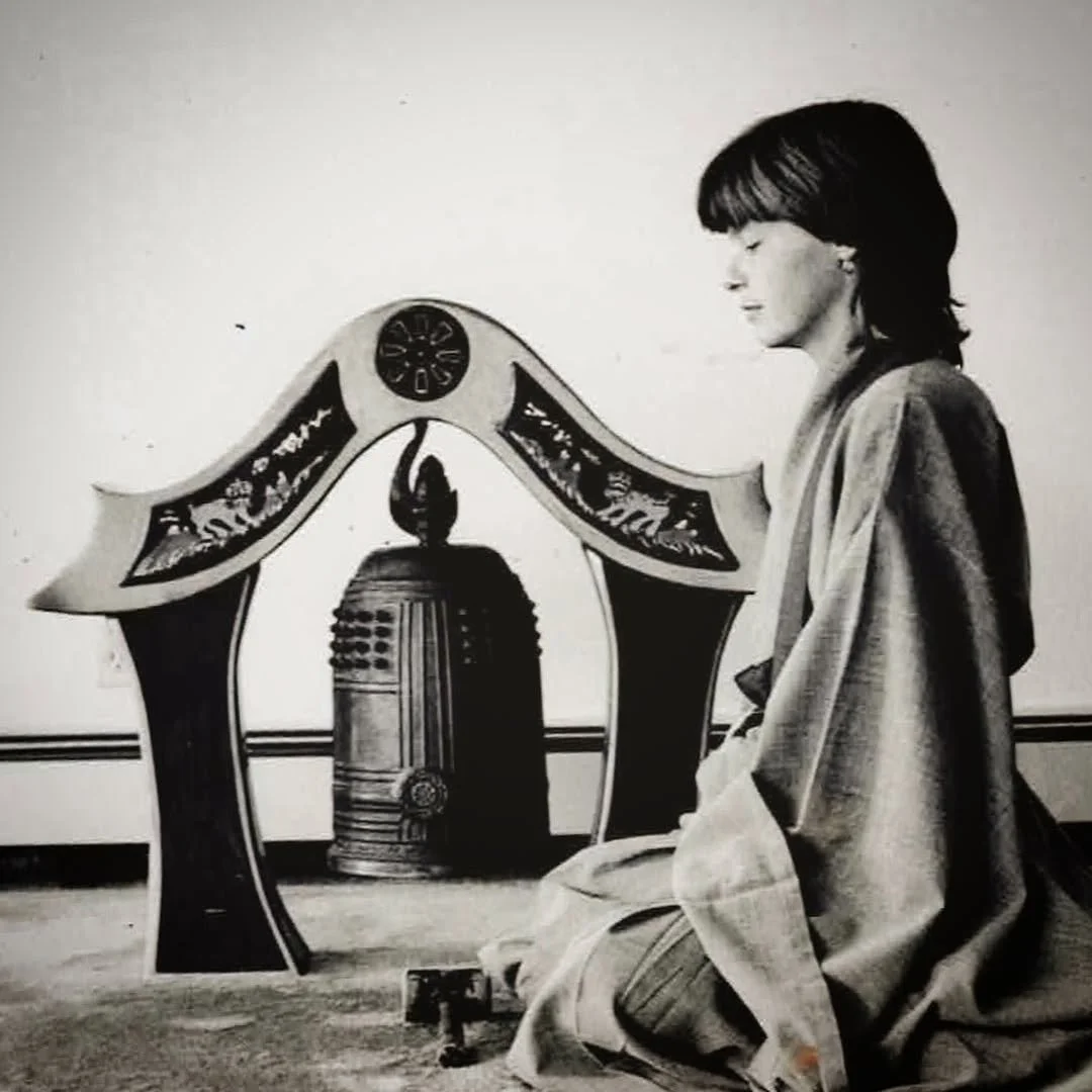 A woman with short dark hair wearing traditional clothing, kneeling with her eyes closed, in front of a decorative altar with a large candle and ornate details, in black and white.