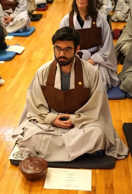 A man with glasses and a beard, sitting cross-legged on a meditation cushion, wearing monastic robes, in a room with other people sitting in similar postures.