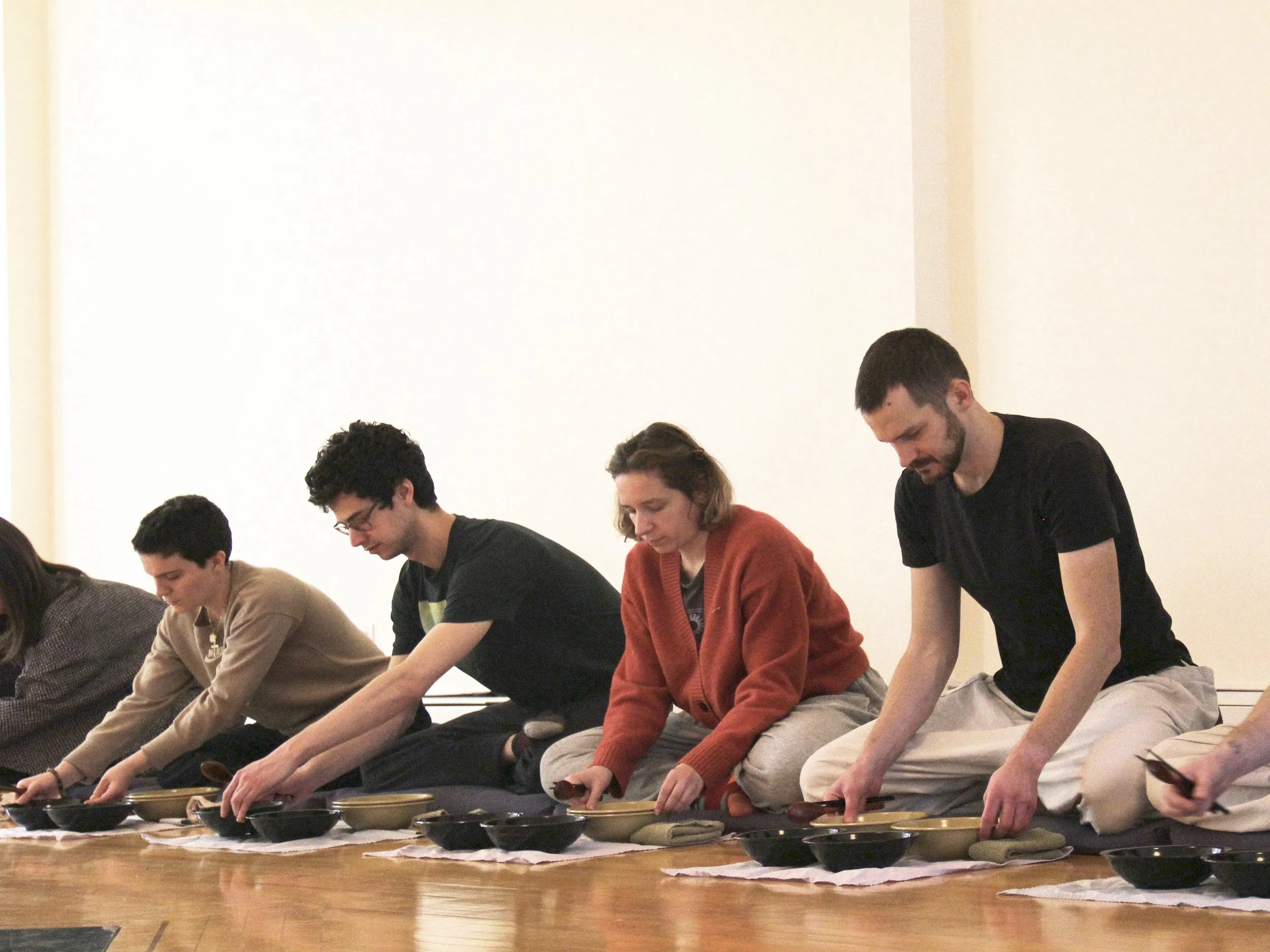 Group of people sitting on the floor with dishes in front of them, possibly participating in a meditation or cultural activity.