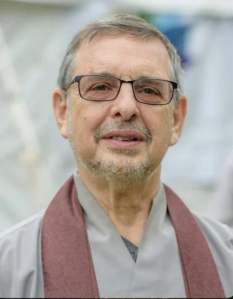 A middle-aged man with gray hair, beard, glasses, wearing a gray shirt and maroon stole, standing outdoors.