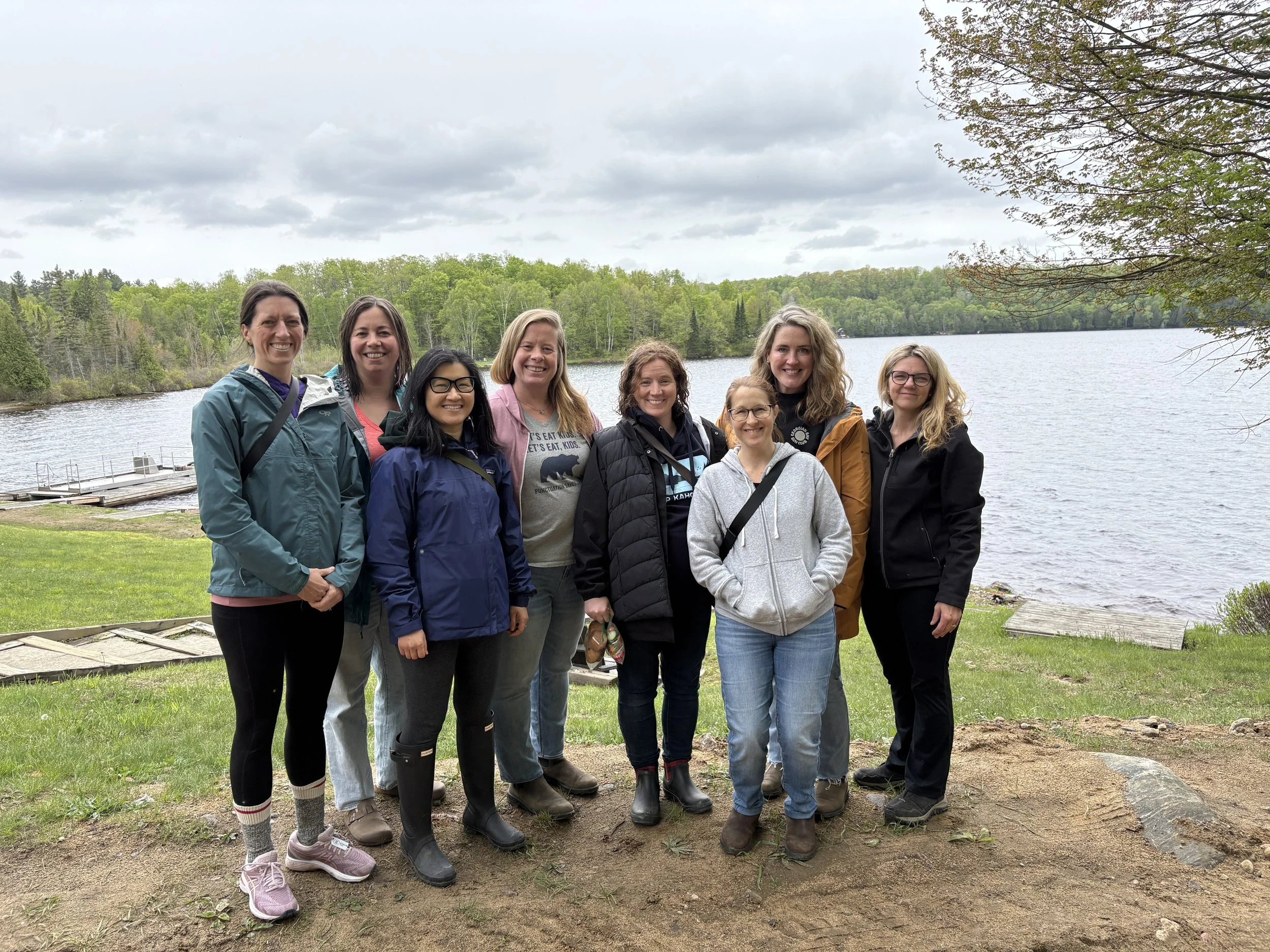 Group of nine women standing outdoors by a lake with trees and cloudy sky in the background.