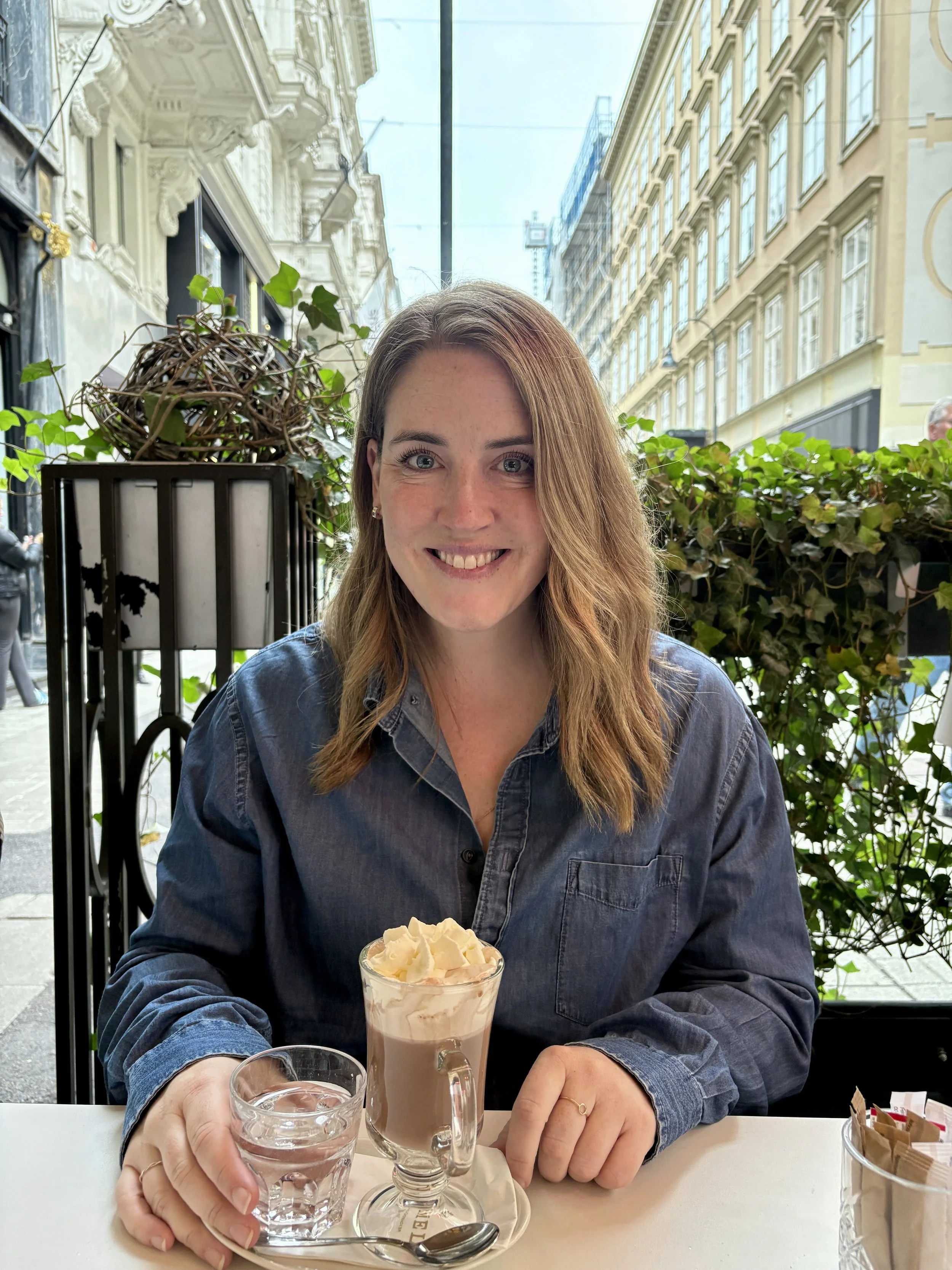 A woman with long brown hair and a denim shirt is smiling at the camera while sitting at a table in a cafe. She has a glass of iced coffee topped with whipped cream in front of her, and she is holding a glass of water. On the table, there is a spoon, a small saucer, and a basket with sugar packets. The cafe has large windows with reflections of buildings outside and decorative white molding on the wall.