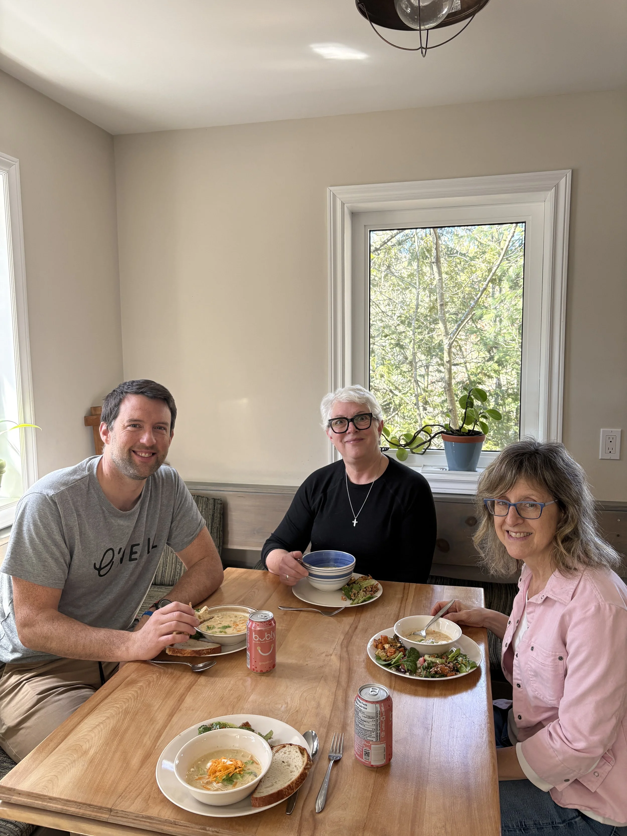 Three people sitting at a wooden dining table enjoying a meal. The man on the left is smiling, holding a spoon, with a bowl of soup and a slice of bread in front of him. The woman in the middle has gray hair, wearing glasses and a black top, holding a bowl of soup and a plate of salad. The woman on the right has curly gray hair, wearing glasses and a pink shirt, also with a bowl of soup and salad. There are soda cans, utensils, and additional bowls of soup and salad on the table. The background features a window with natural light and a leafy view outside.