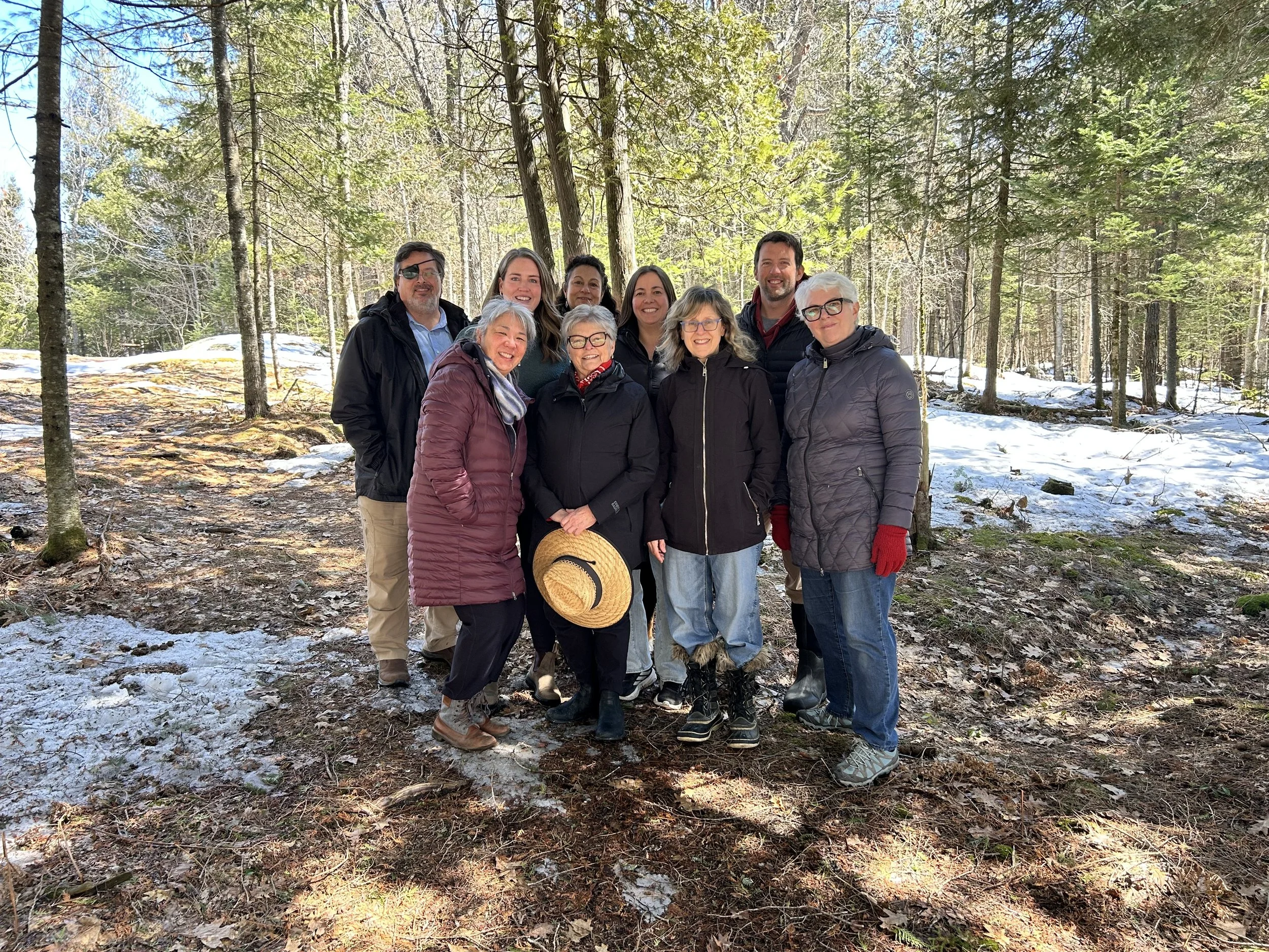 Group of nine adults standing together outdoors in a forest with trees and snow on the ground, dressed in winter clothing and smiling at the camera.