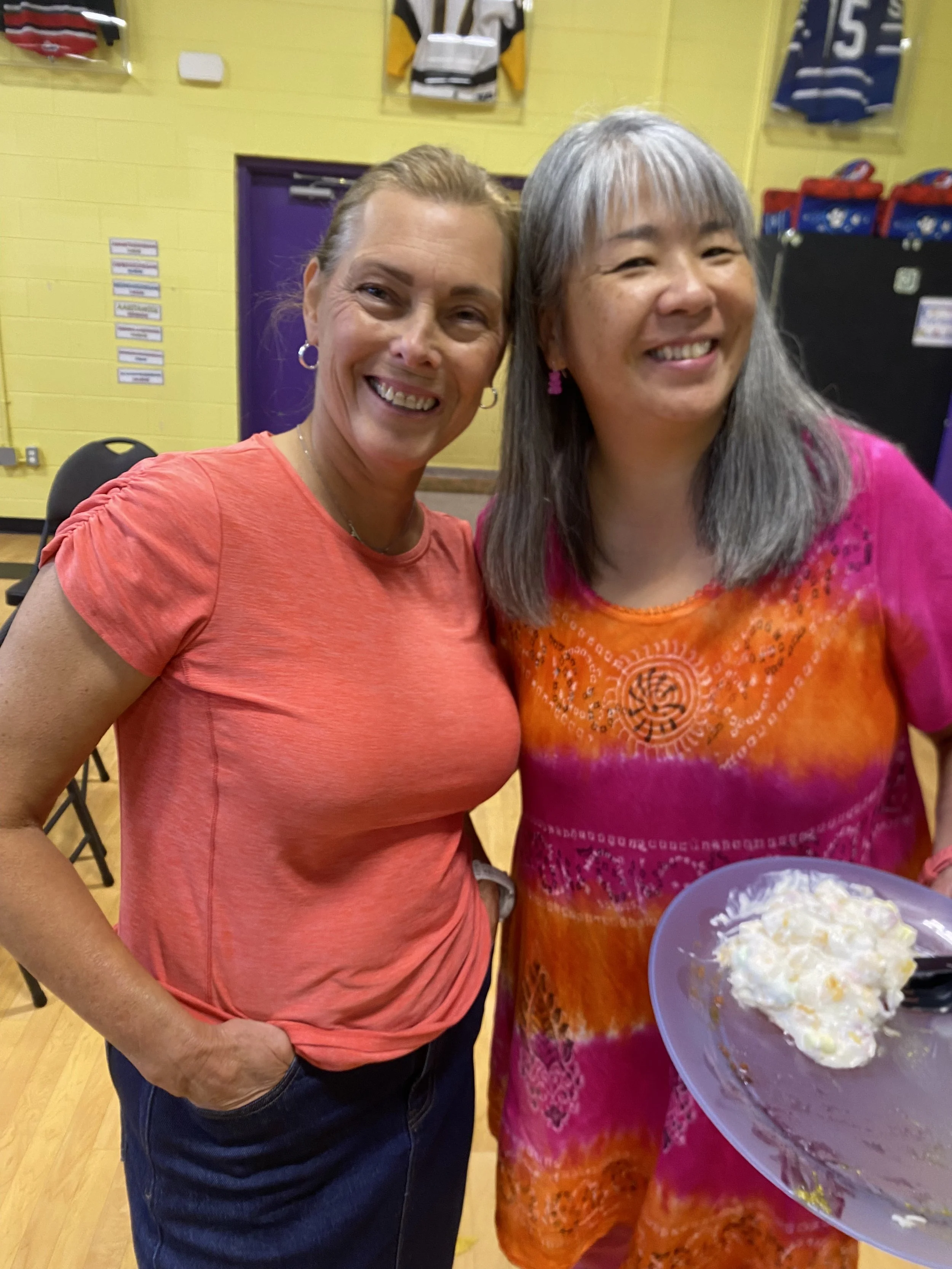 Two women smiling, one holding a plate of cake at a colorful indoor event, with sports jerseys hanging on a yellow wall in the background.