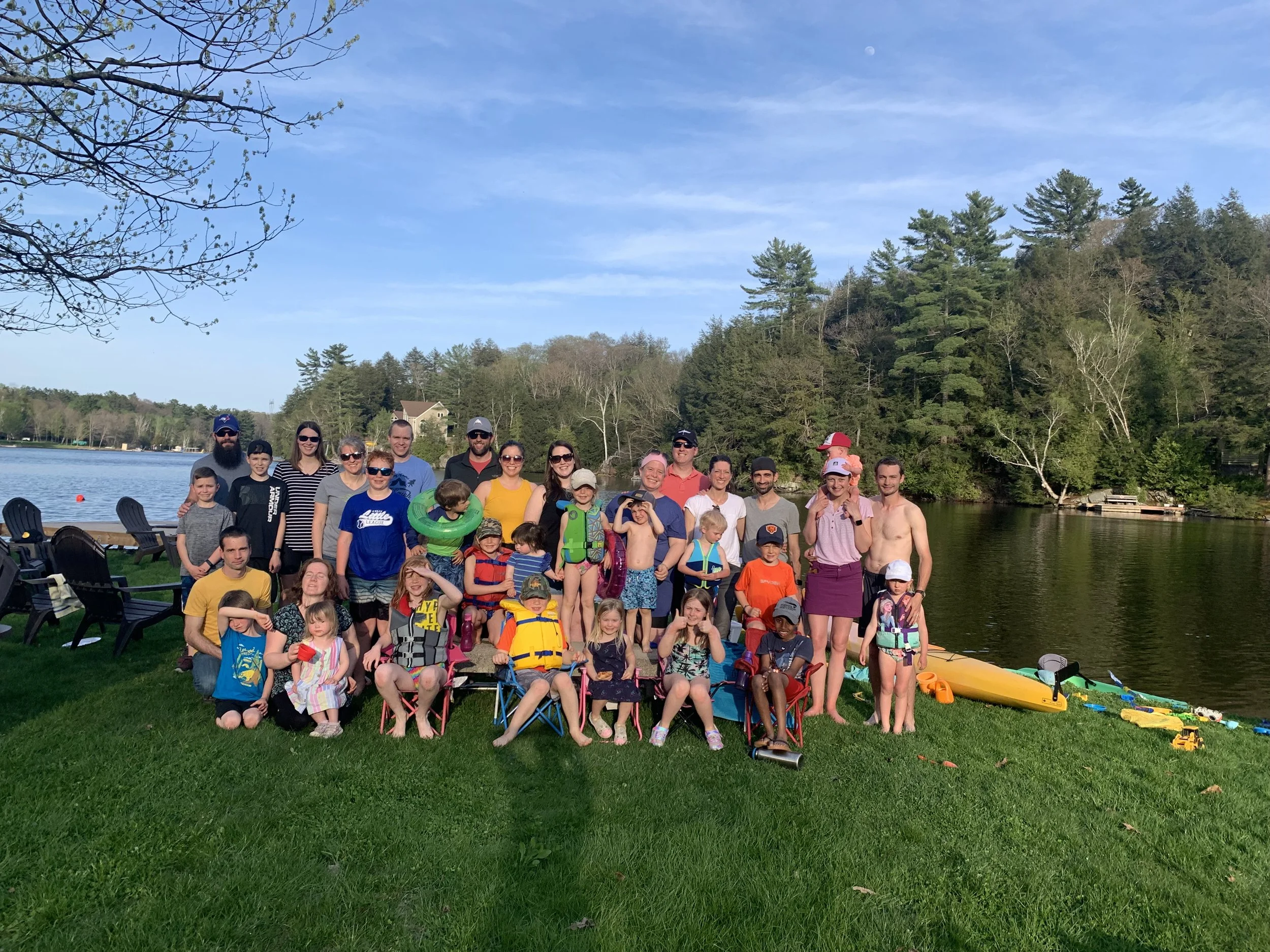 A large group of children and adults gathered outdoors near a body of water, with trees and a clear sky in the background, some children wearing life jackets and holding water toys.