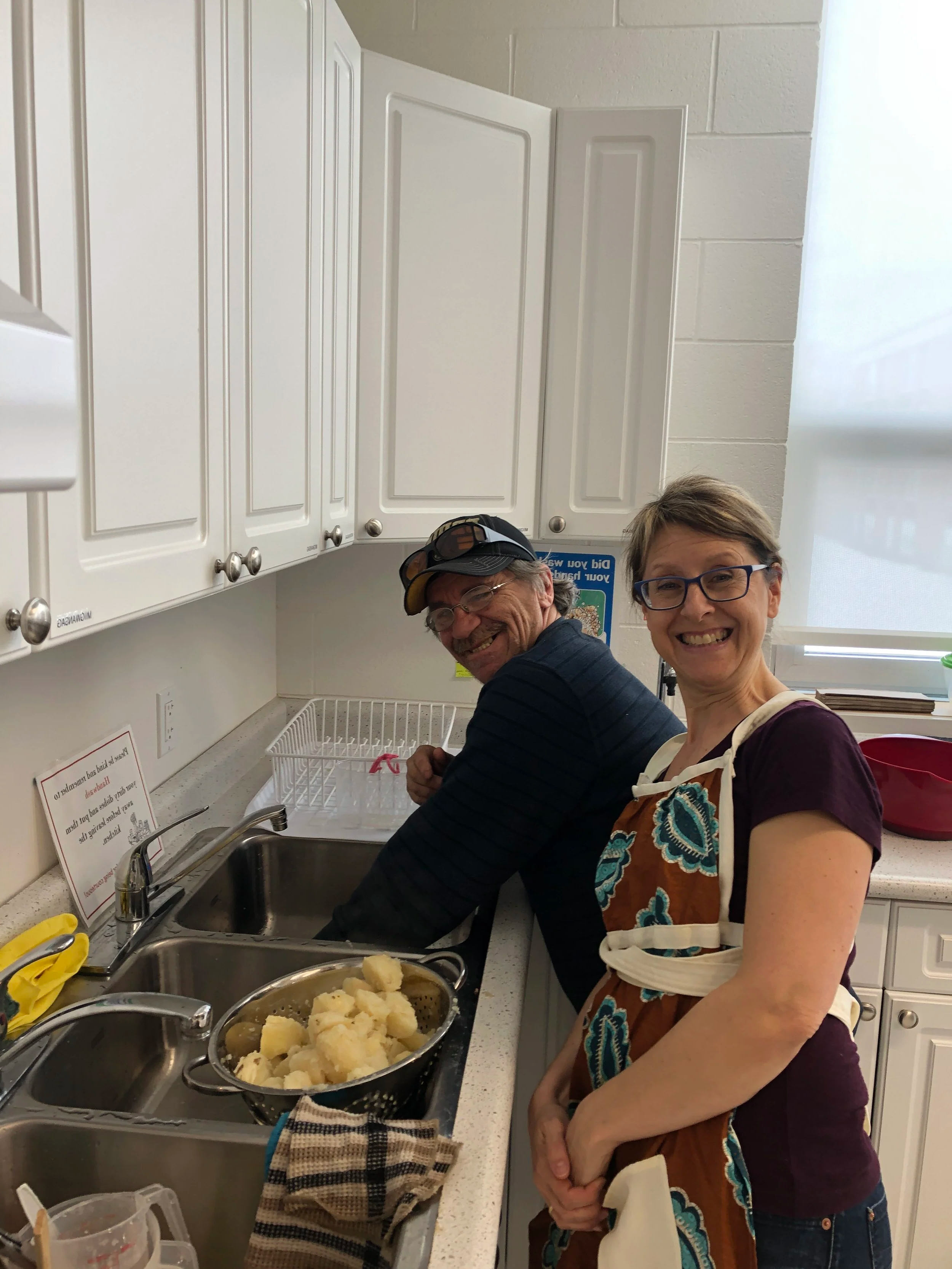Two people smiling and standing at a kitchen sink with dishes, potatoes, and kitchen cabinets visible in the background.
