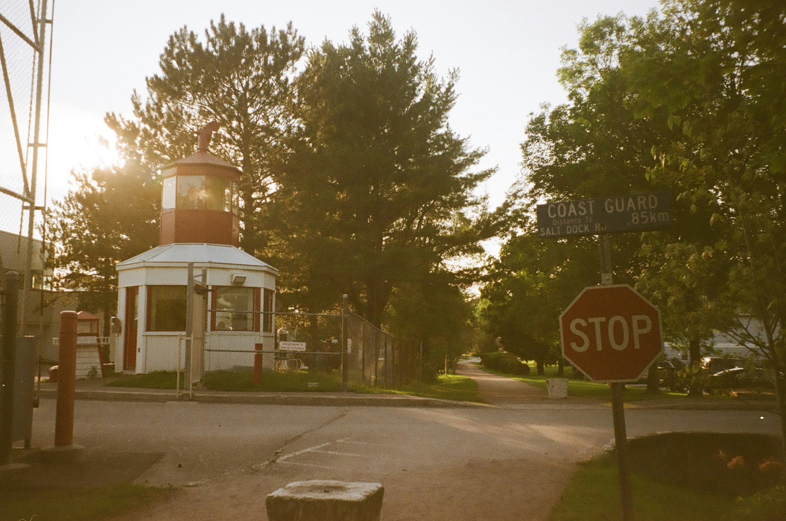 A security checkpoint with a small guard booth, a stop sign, a fence, and a sign indicating the Coast Guard is 0.85 km away, with trees and the sun in the background.