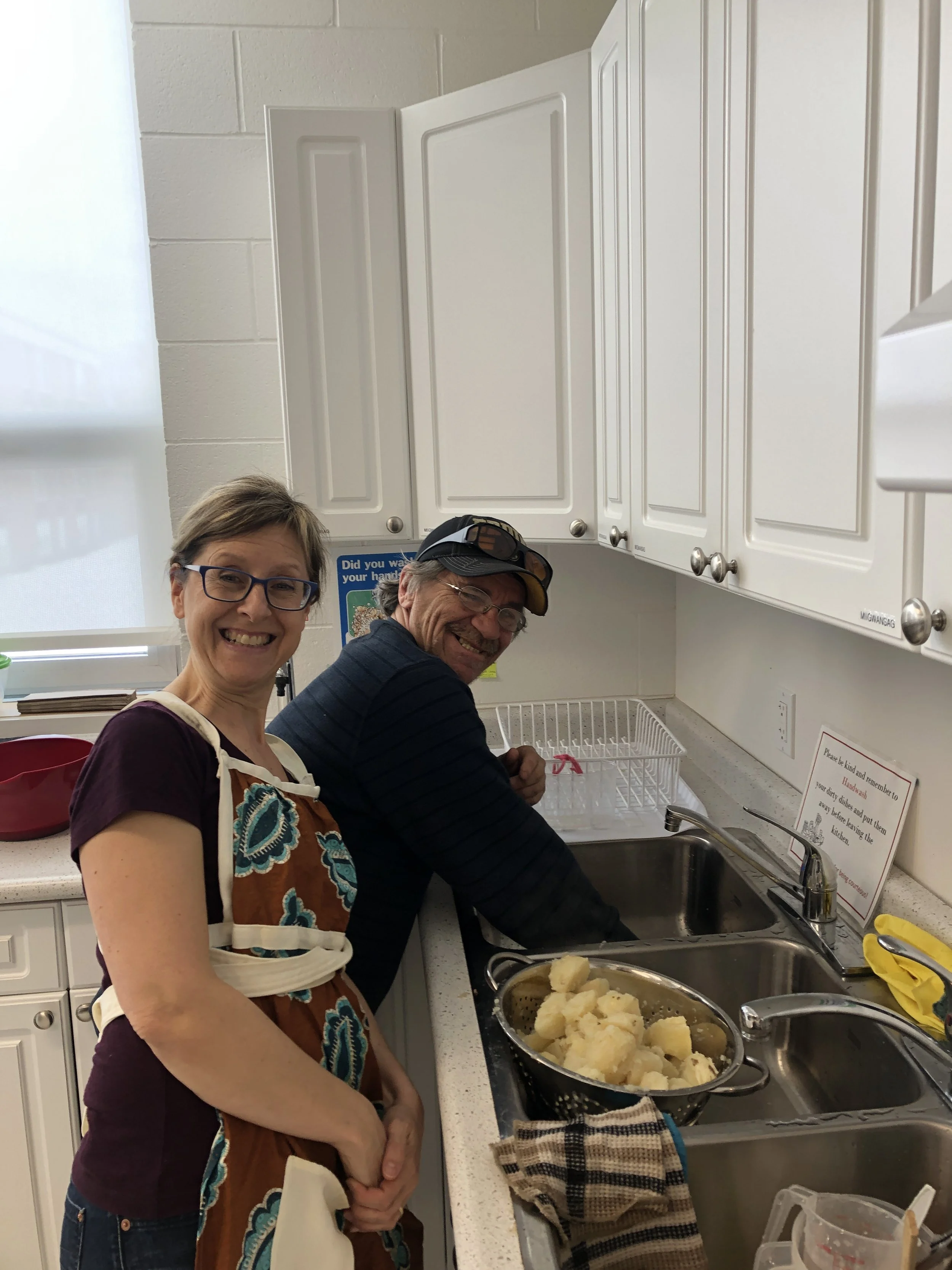 Two smiling people, a woman and a man, cooking potatoes in a kitchen. The woman is wearing a colorful apron and glasses, while the man is wearing a cap and glasses. They are standing by a sink with a pot of cooked potatoes and a towel nearby.