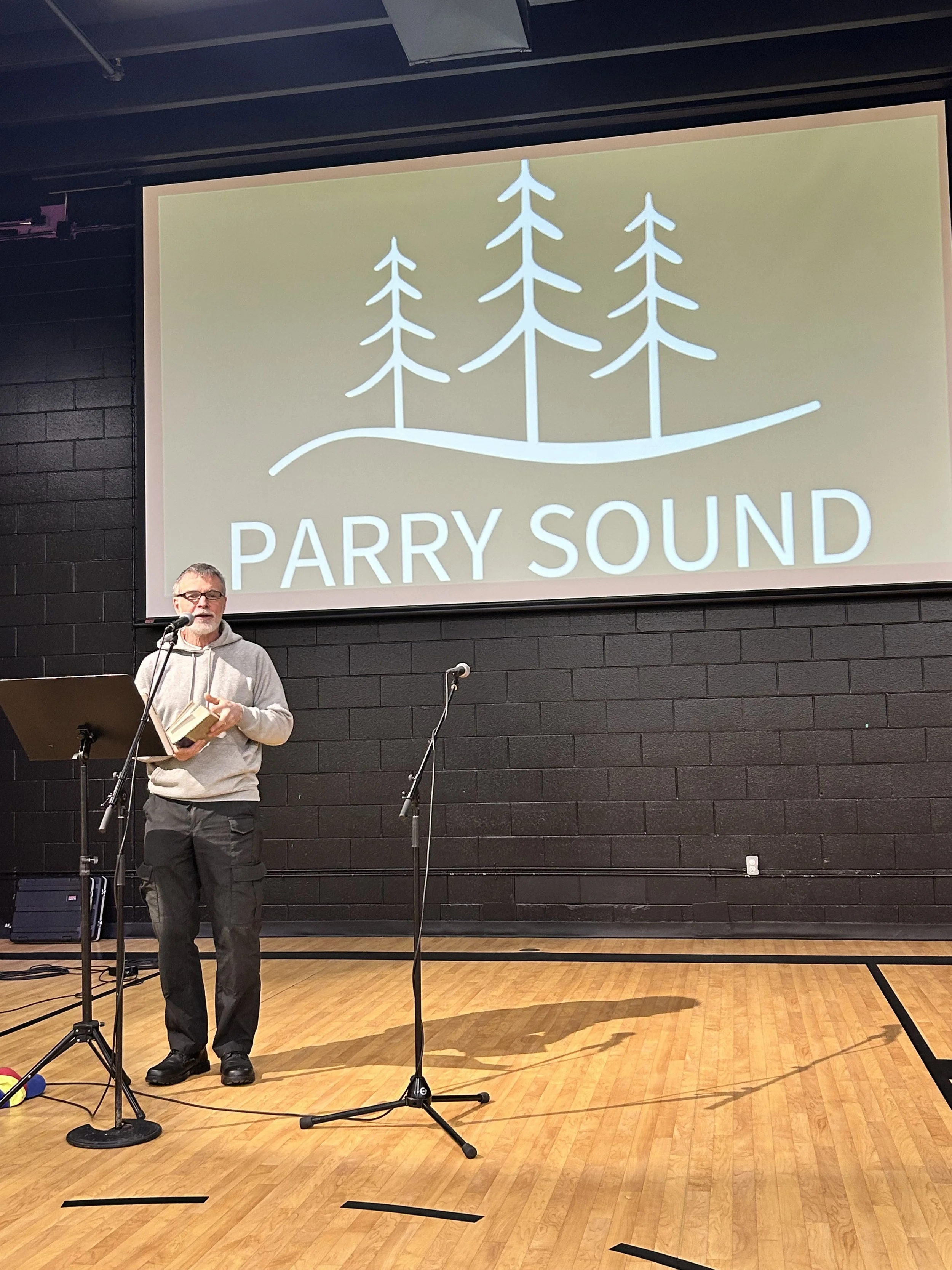 A man holding a book and speaking into a microphone on a stage with a large screen behind him displaying the logo 'PARRY SOUND' and three stylized trees.