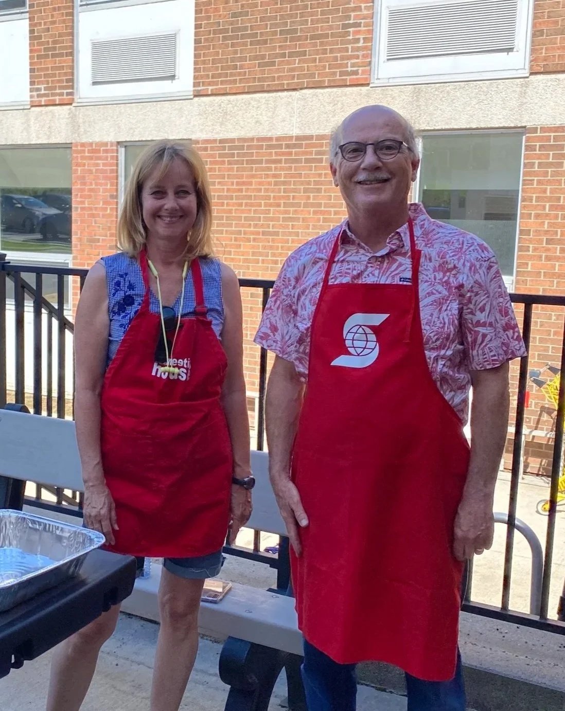 A woman and a man standing outdoors on a patio, both wearing red aprons. The woman has blonde hair, is wearing a sleeveless top, and appears to be holding a pair of sunglasses. The man has thinning hair and glasses, wearing a patterned short-sleeve shirt. They are smiling and standing in front of a black railing with a building with brick walls and windows in the background.