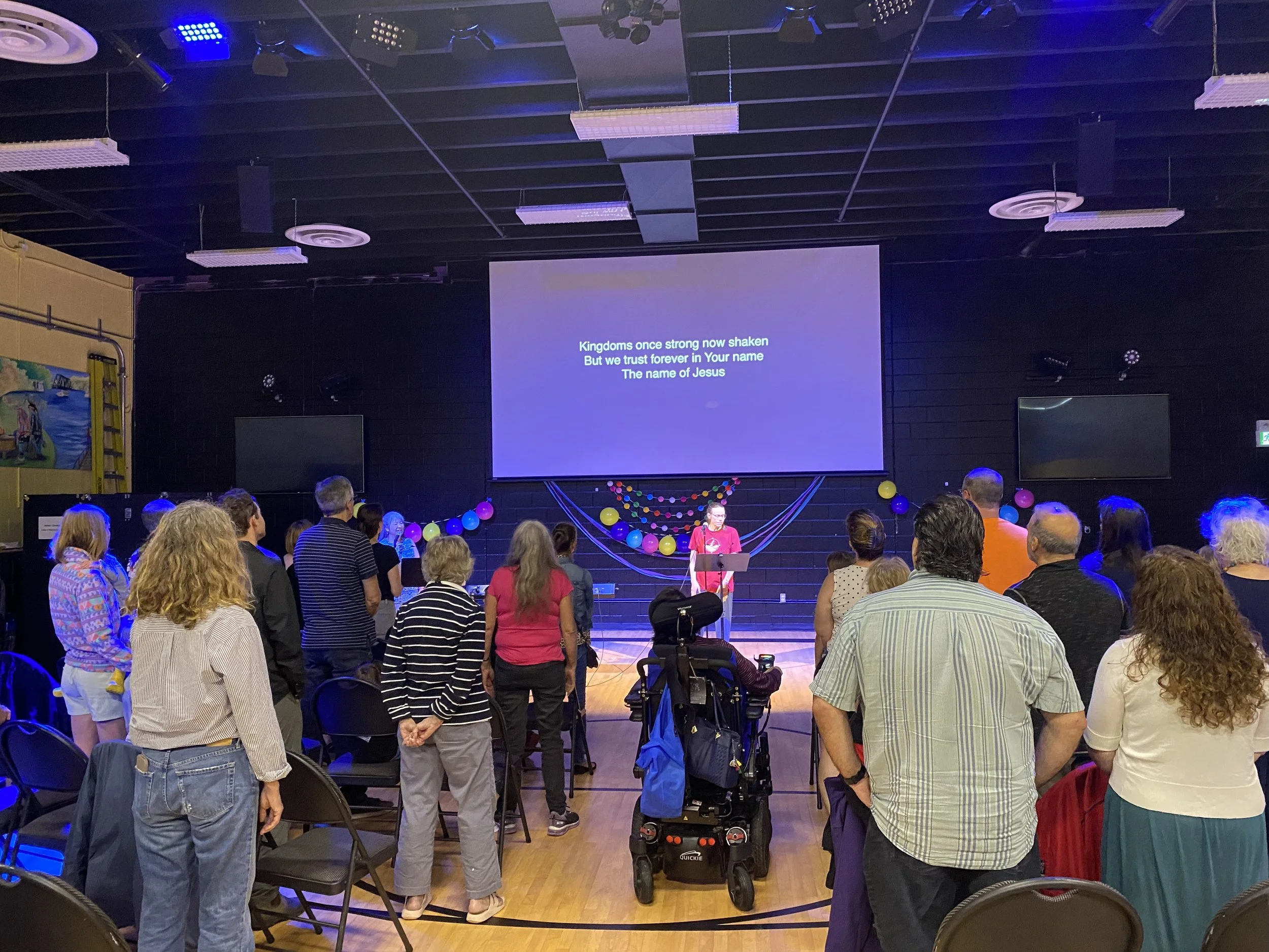 A group of people gathered in a room with a stage showing lyrics on a large screen. The room is decorated with balloons and streamers, and the audience is standing and facing the stage where a person is singing or speaking.