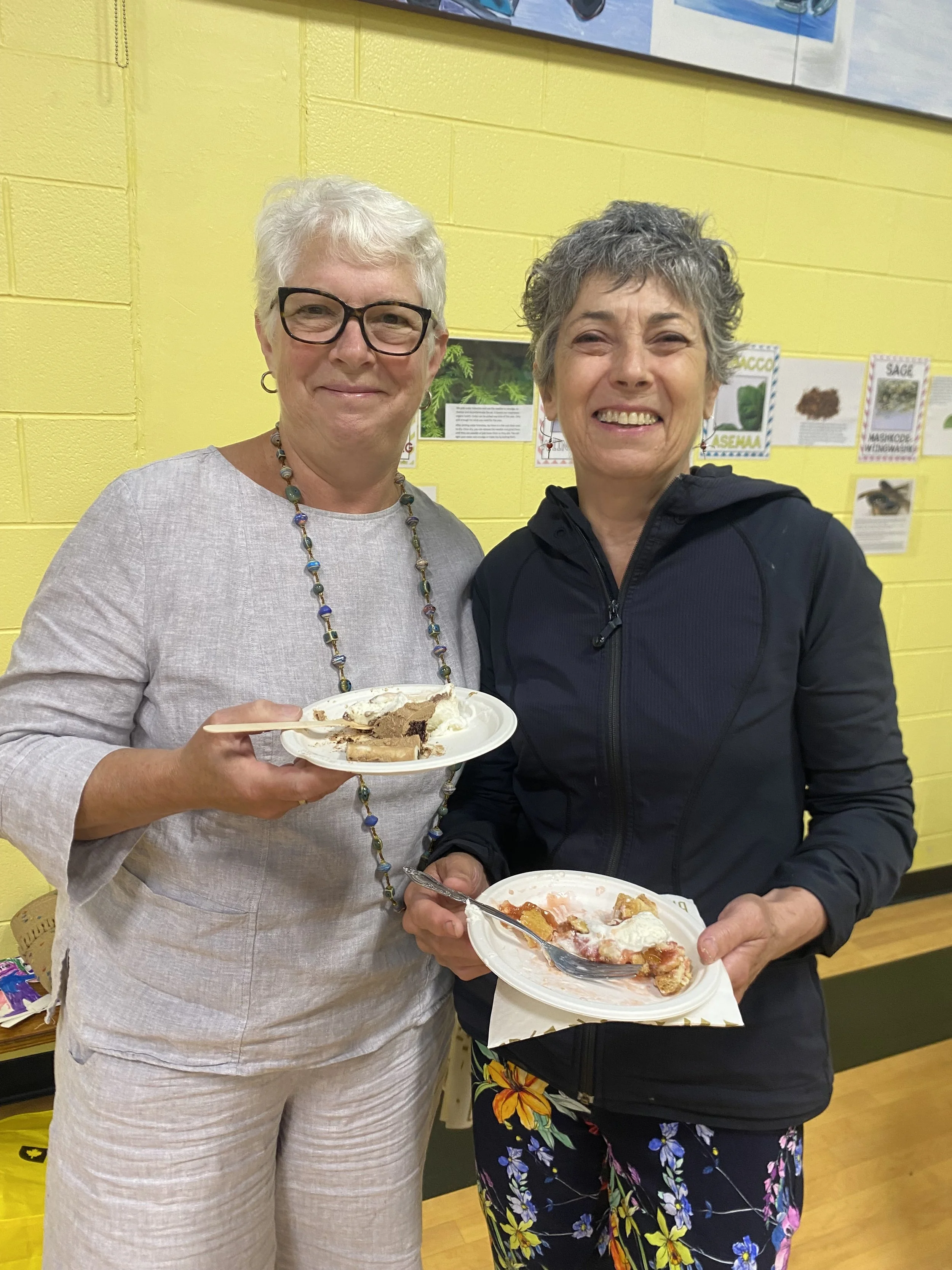 Two women smiling and holding plates of food, standing in front of a yellow wall with educational posters about herbs and plants.
