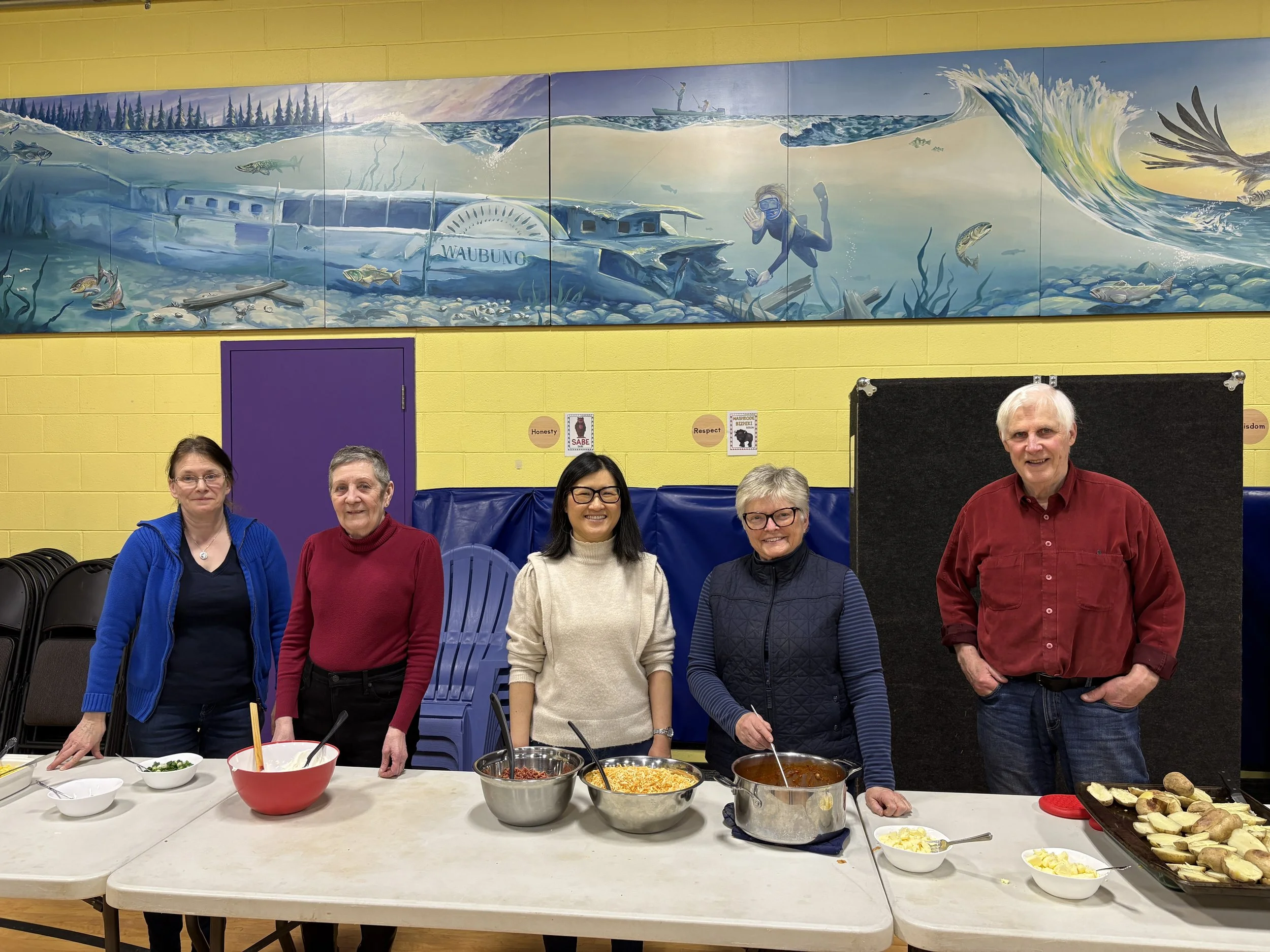 Group of five people standing behind a table with bowls and food, in a room with yellow walls and a mural of a sunlit ocean scene with fish, underwater ruins, and a swimmer.