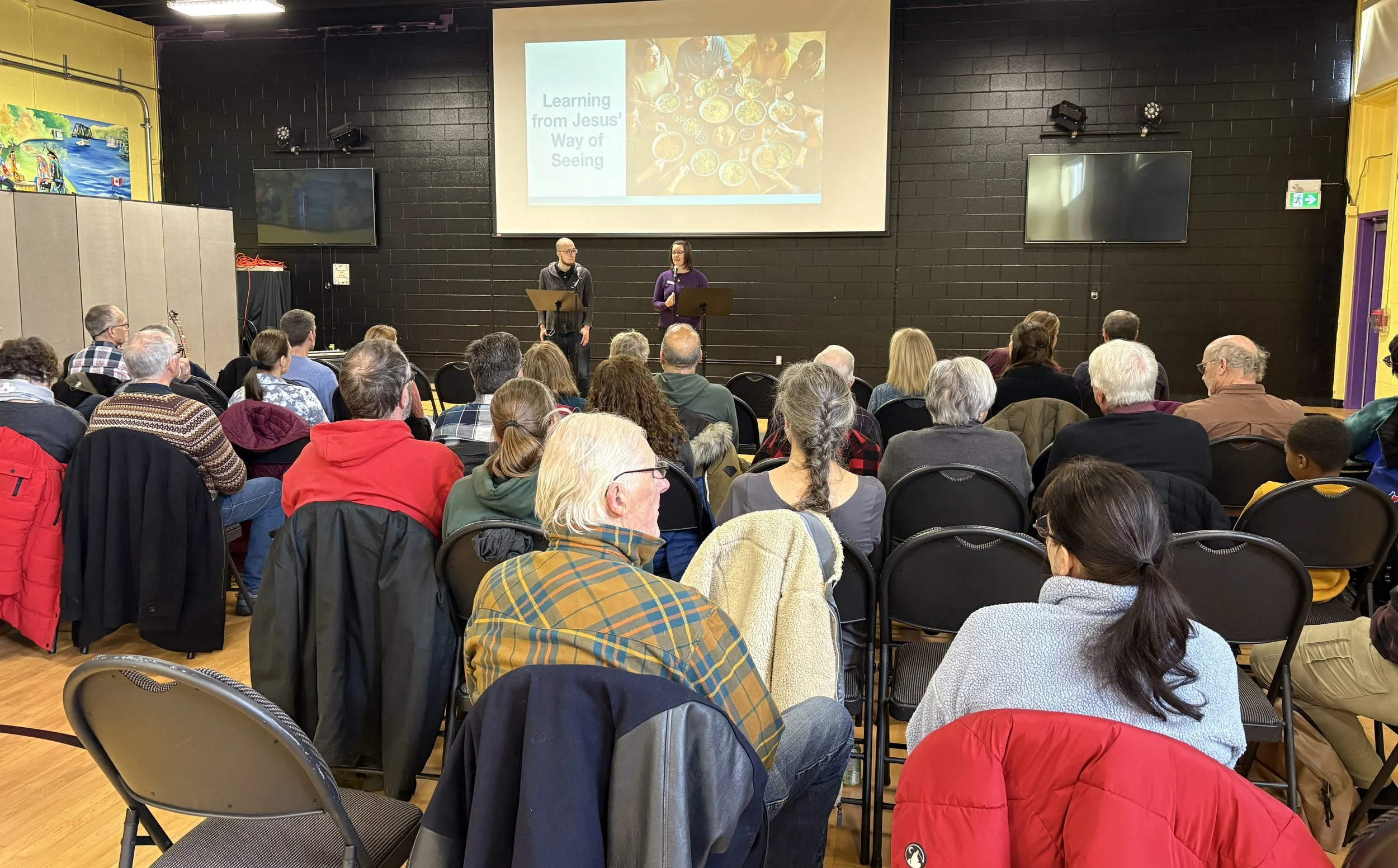 A crowd of people seated in chairs listening to two speakers standing on a stage in front of a large screen. The screen displays a presentation slide with the text 'Learning from Jesus' Way of Seeing' and an image of several people sharing a meal.