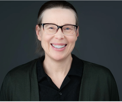 A woman with glasses and light-colored hair pulled back, smiling against a dark background.