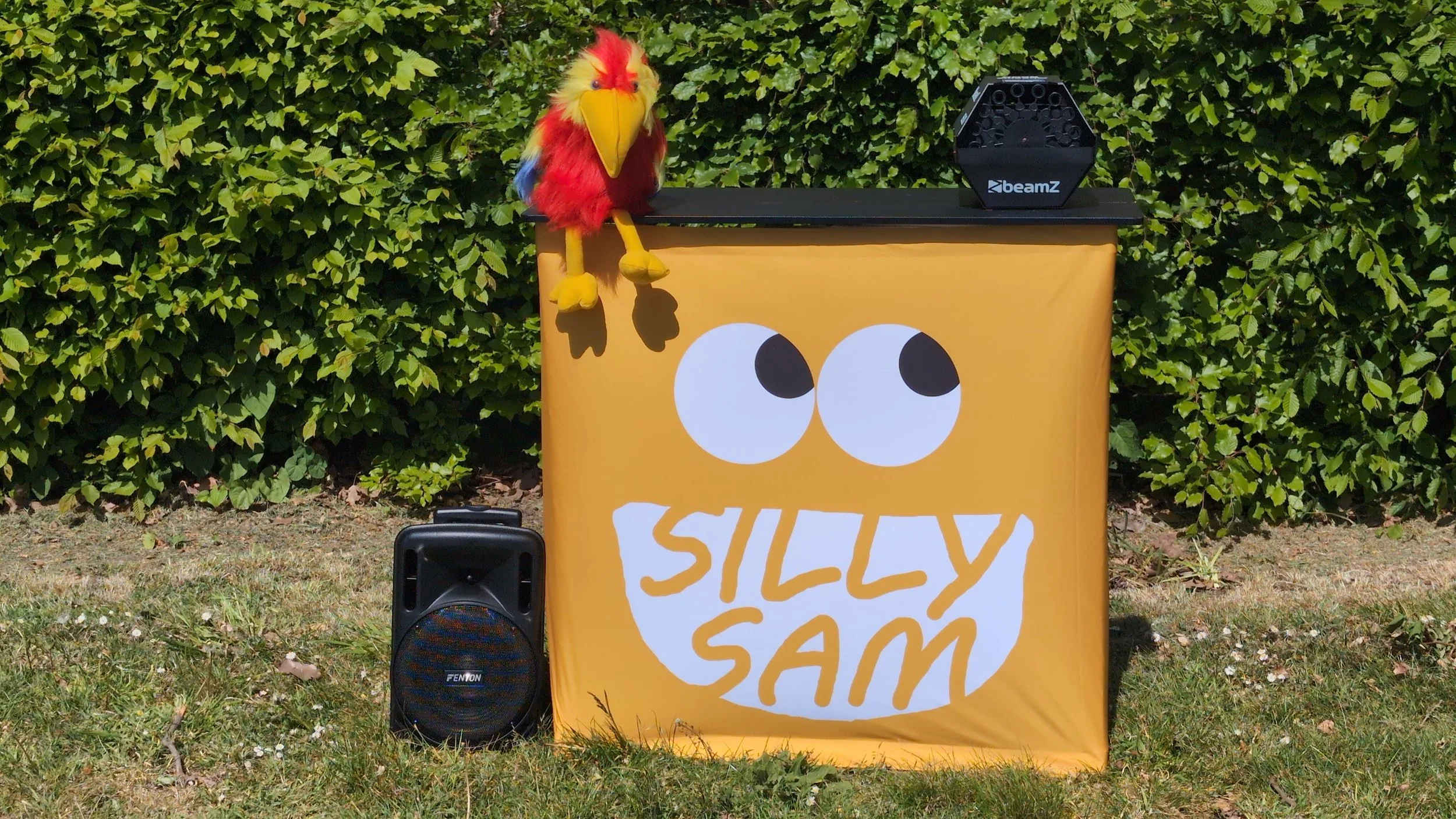 A yellow and red puppet bird with a large beak sitting on a black table with a speaker next to it. The table has a cartoon-like face with large eyes and the words 'SILLY SAM' on it. The background is green foliage.