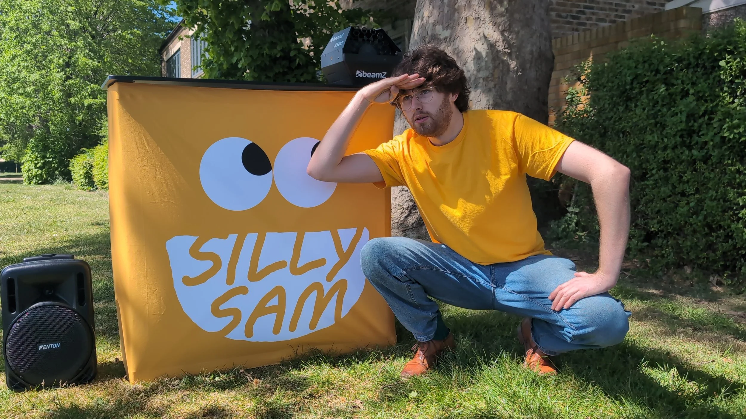 A young man with glasses, curly hair, and a beard is sitting outdoors on grass, wearing a yellow t-shirt and jeans, giving a salute pose with one hand while other hand is on his knee. Behind him is a yellow banner with white cartoon eyes and the words "Silly Sam." A black speaker is on the ground nearby, and green trees and bushes are in the background.
