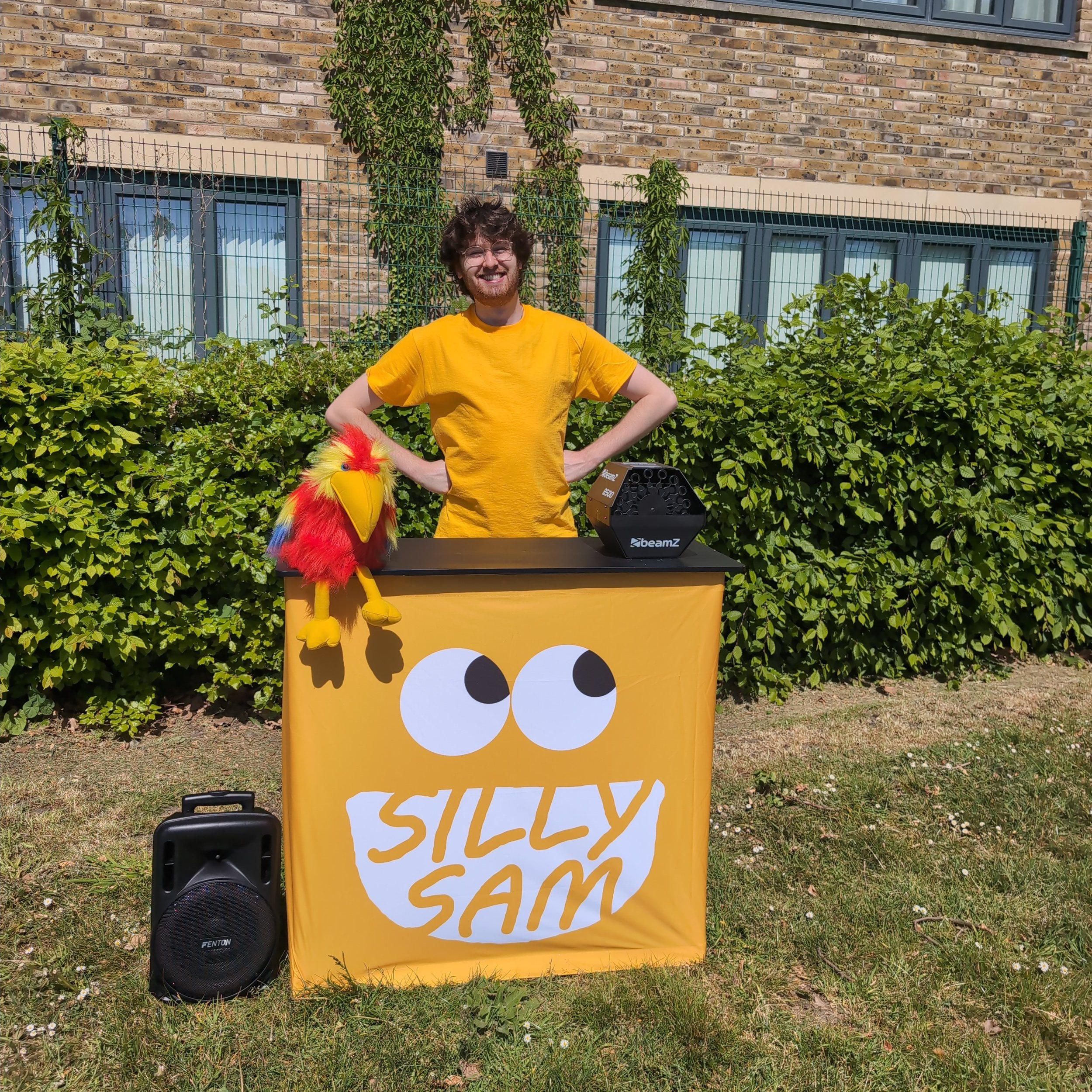 A man in a yellow T-shirt standing behind a yellow booth with a face design, outdoors with green bushes and a brick building in the background. A colorful puppet and a speaker are on the table.