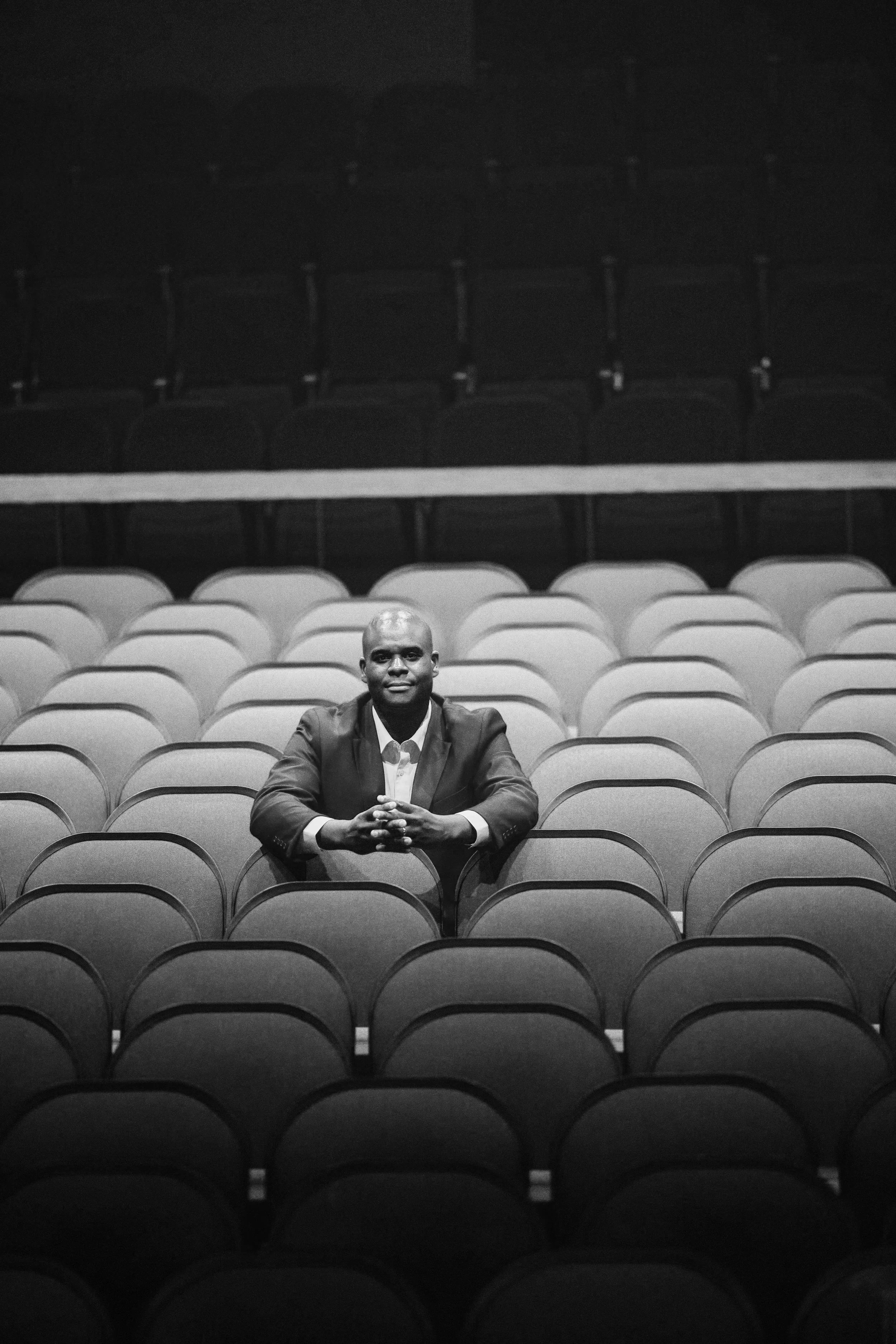 A man in a suit sitting alone in a large, empty auditorium with multiple rows of empty chairs, looking directly at the camera with a confident expression.